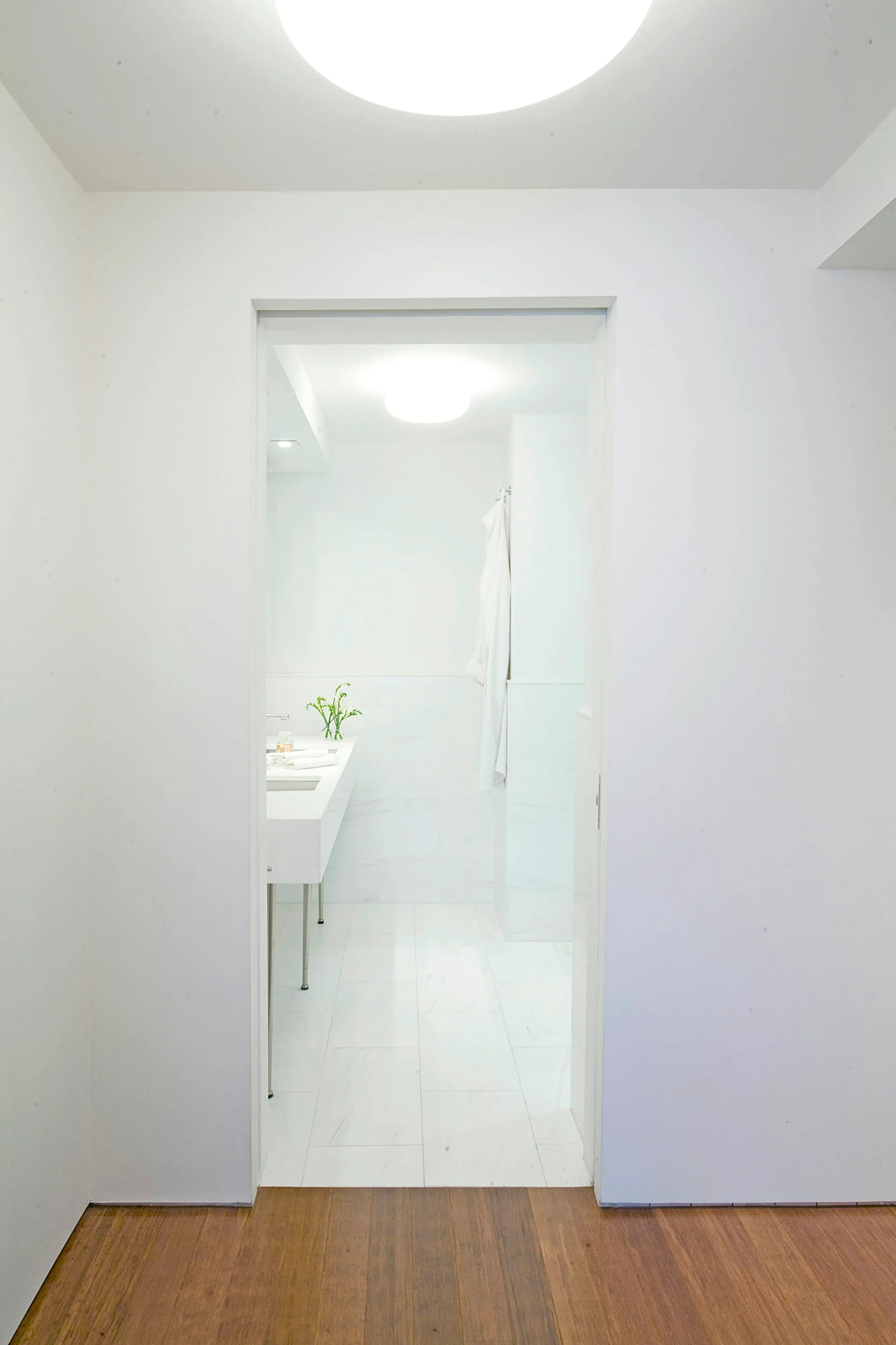 Minimalist bathroom entry with frosted glass sliding door, white walls, warm wood flooring, and modern ceiling light by Elskop Scholz LLC.