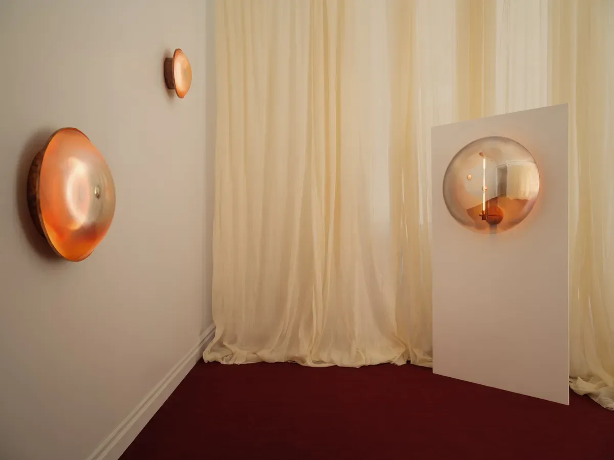 Gallery space with cream curtains, burgundy floor, and three copper-toned circular wall-mounted light installations by Emma Scully Gallery.