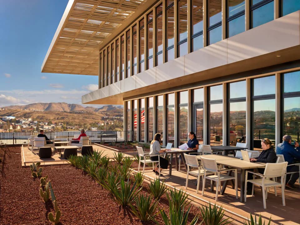 Modern multi-story building with expansive glazing, wooden ceiling elements, and terrace with mountain views, featuring contemporary furniture and landscaping by Ennead Architects.