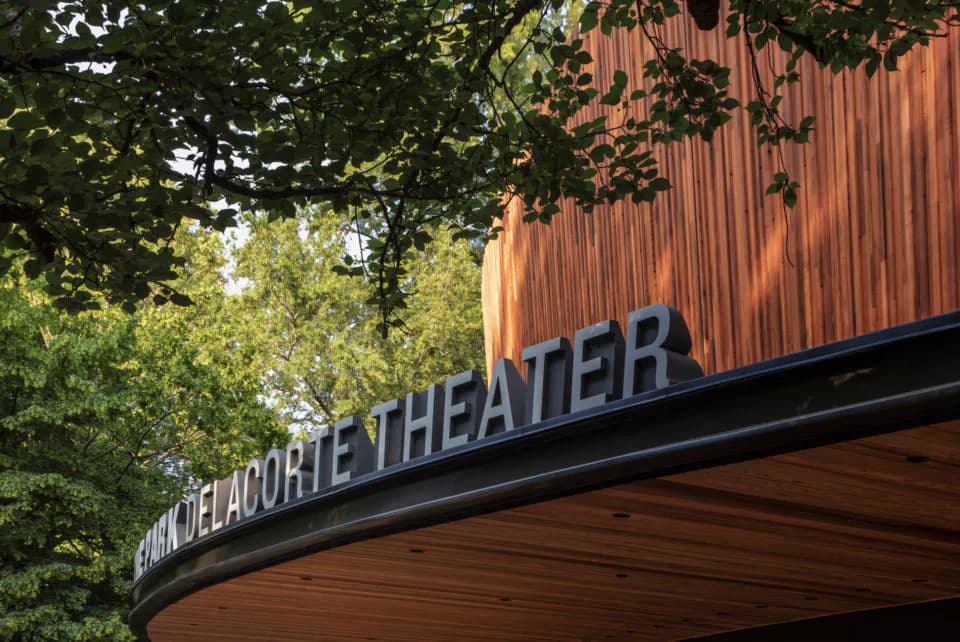 Modern theater marquee with curved wooden soffit and signage integrated into the architecture, surrounded by mature trees, by Ennead Architects.