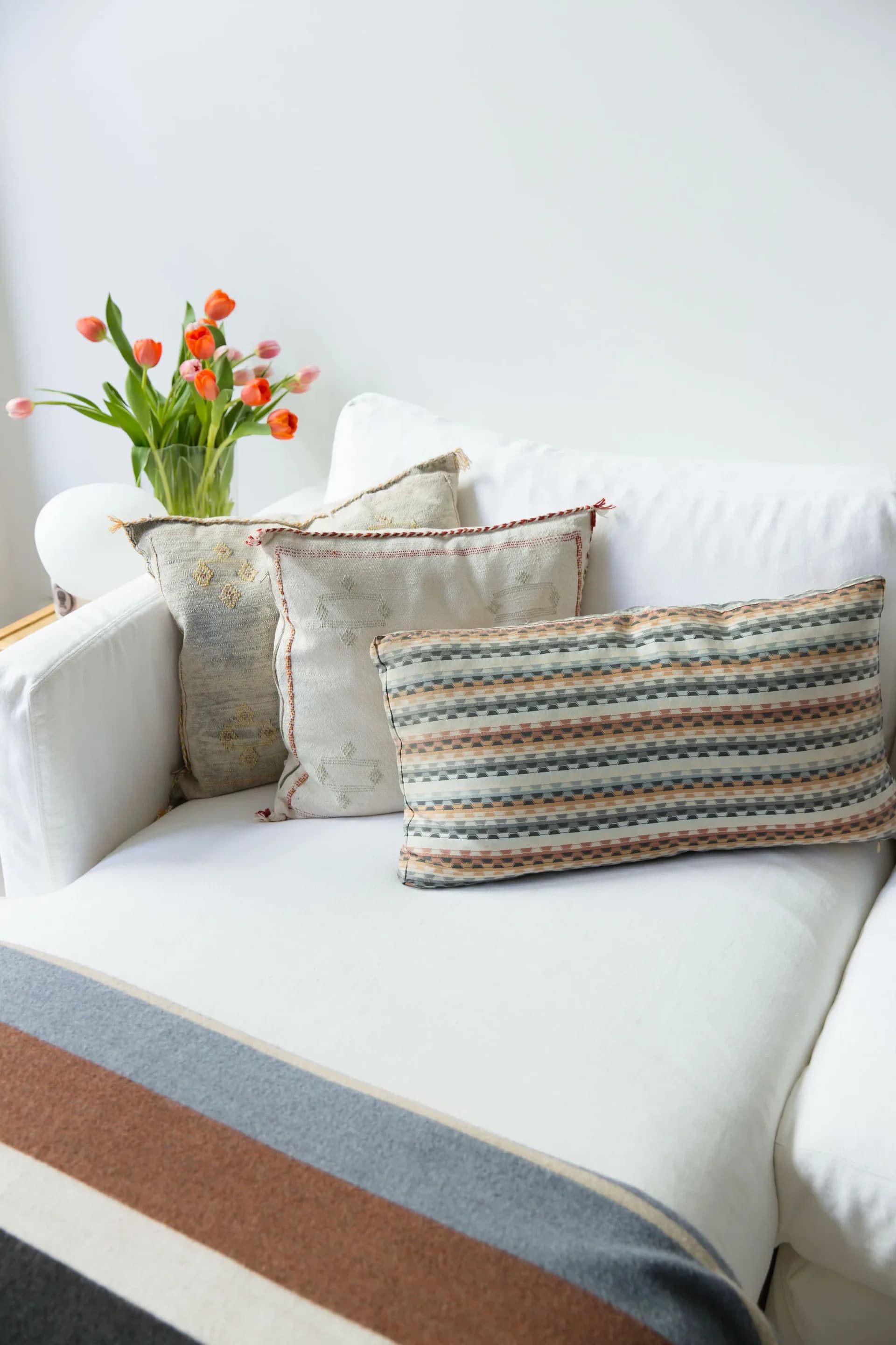 White upholstered sofa styled with patterned and textured throw pillows alongside fresh orange and pink tulips in a clear glass vase by Allison Garcy Interiors.