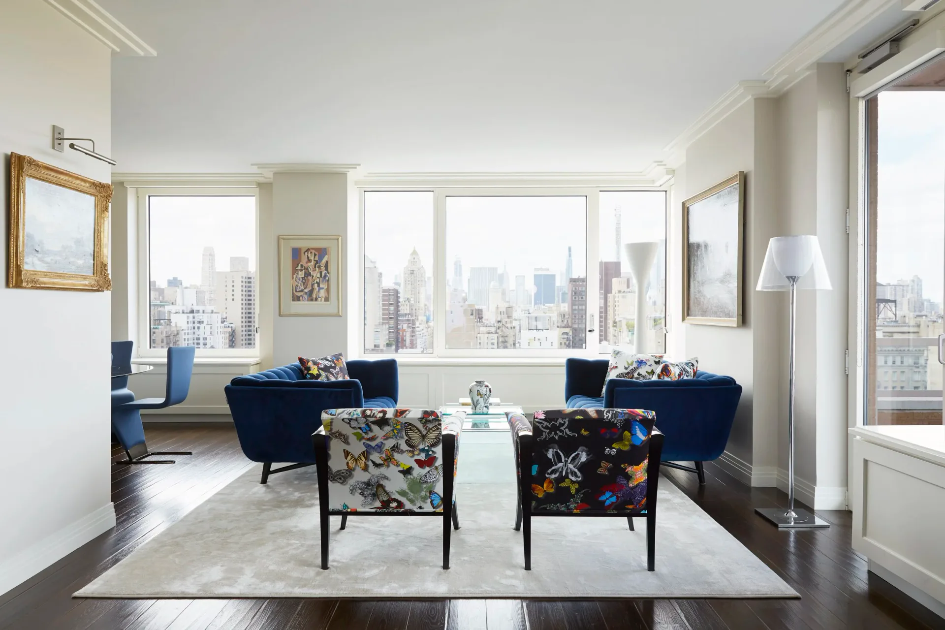 Modern high-rise living room with floor-to-ceiling windows overlooking a city skyline, navy blue armchairs, patterned accent chairs, and light wood flooring by Allison Garcy Interiors.