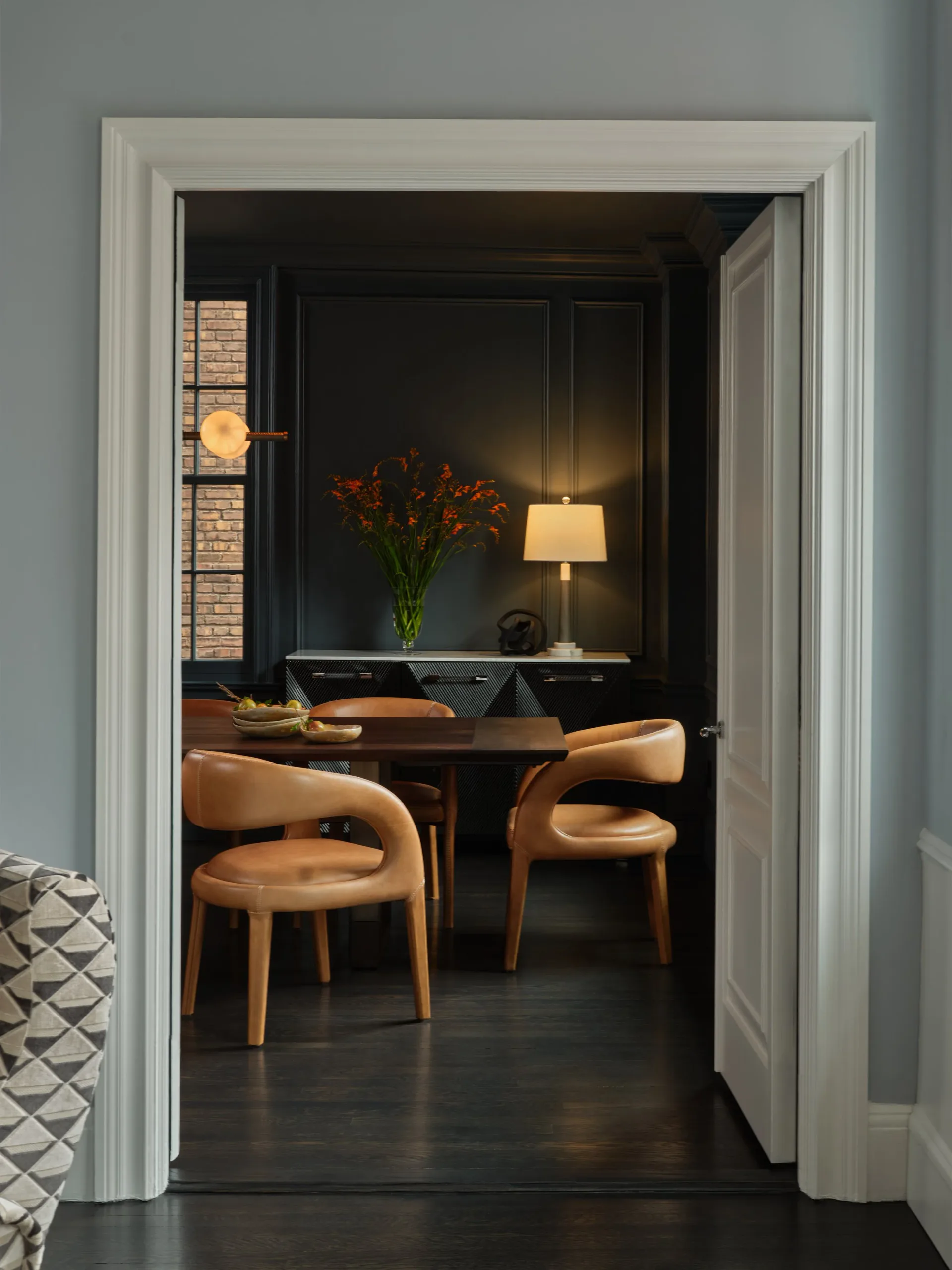 Modern dining room with cognac leather chairs, dark wood table, and dramatic black walls accented by warm lighting and floral arrangement by Allison Garcy Interiors.