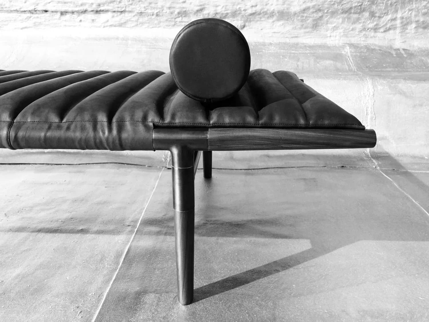Minimalist black bench with quilted seat and spherical headrest casting sharp shadows on polished concrete floor by Erickson Æsthetics.