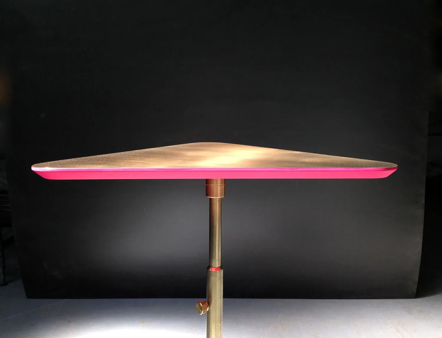A sculptural pendant lamp with a sweeping curved form, illuminated from within with warm golden light and accented with a vibrant pink rim, suspended from a brass pole by Erickson Æsthetics.