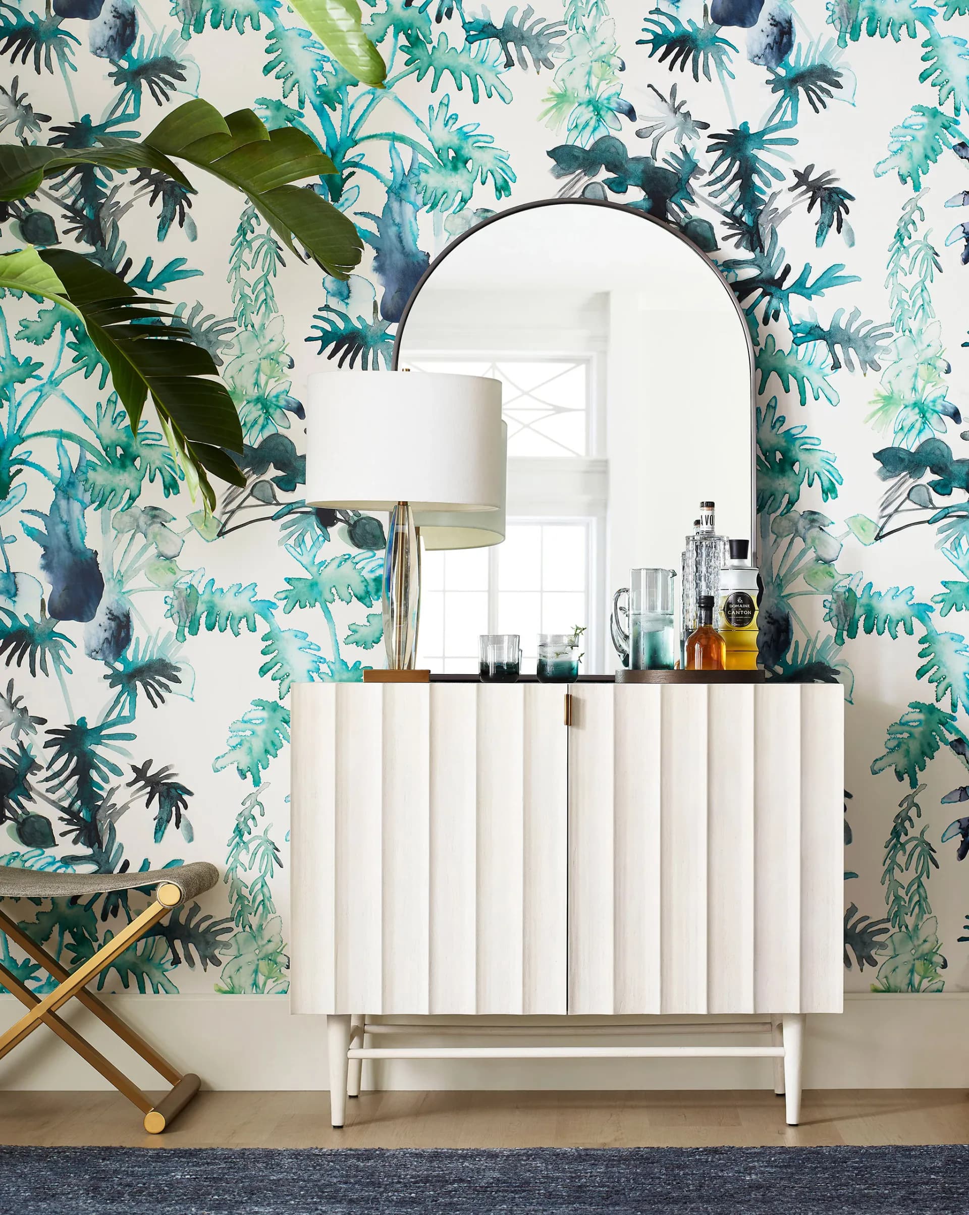 A bedroom vanity with tropical leaf wallpaper in turquoise and green tones, featuring a white ribbed dresser, arched mirror, and warm wood accent chair by Eskayel.