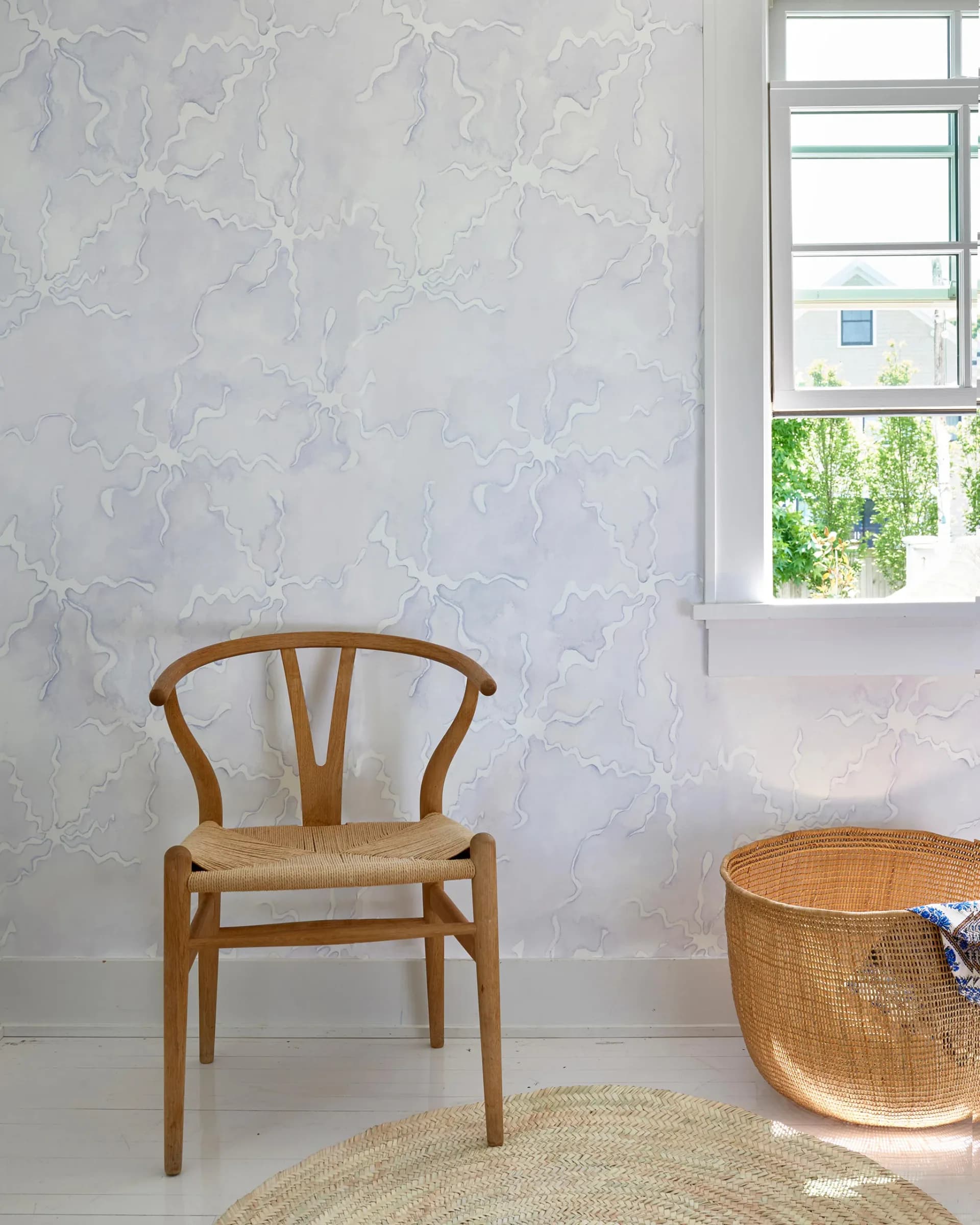 Minimalist interior with textured white wallpaper, wooden wishbone chair with woven seat, woven basket, and natural light from large windows by Eskayel.