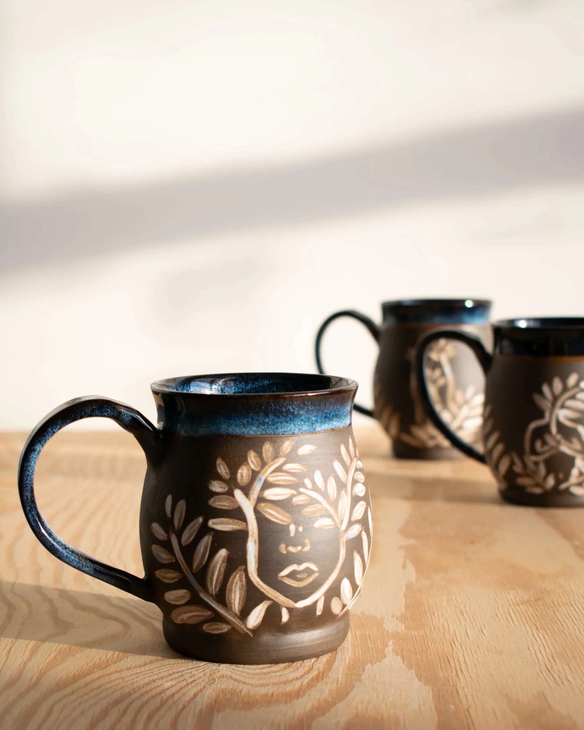 Three handcrafted ceramic mugs with blue-glazed rims and carved leaf-motif faces on dark clay, arranged on light wood by Estelle's Atelier.