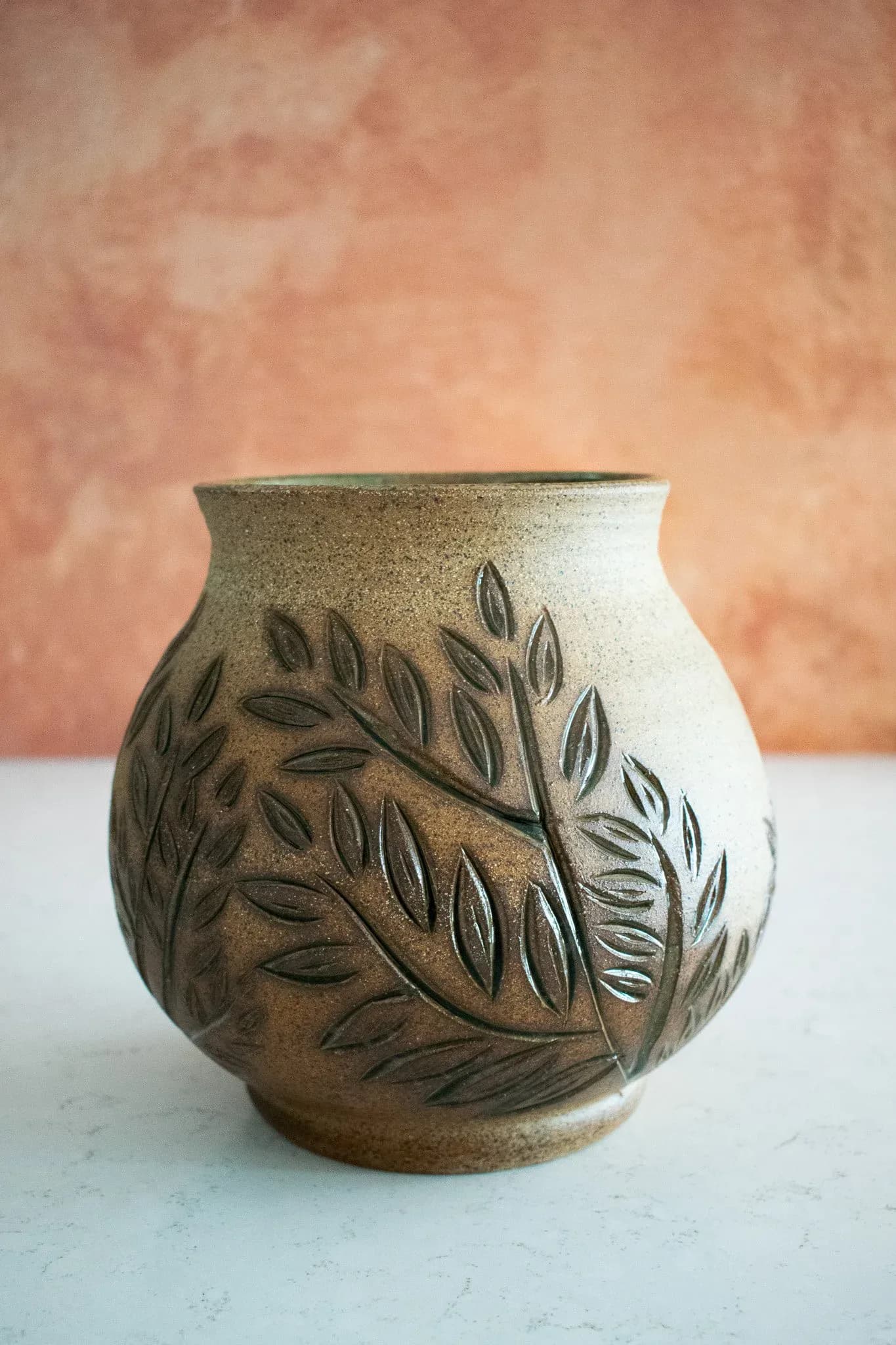 Ceramic vessel with textured cream glaze and carved leaf motifs in dark brown, displayed against a peachy backdrop by Estelle's Atelier.