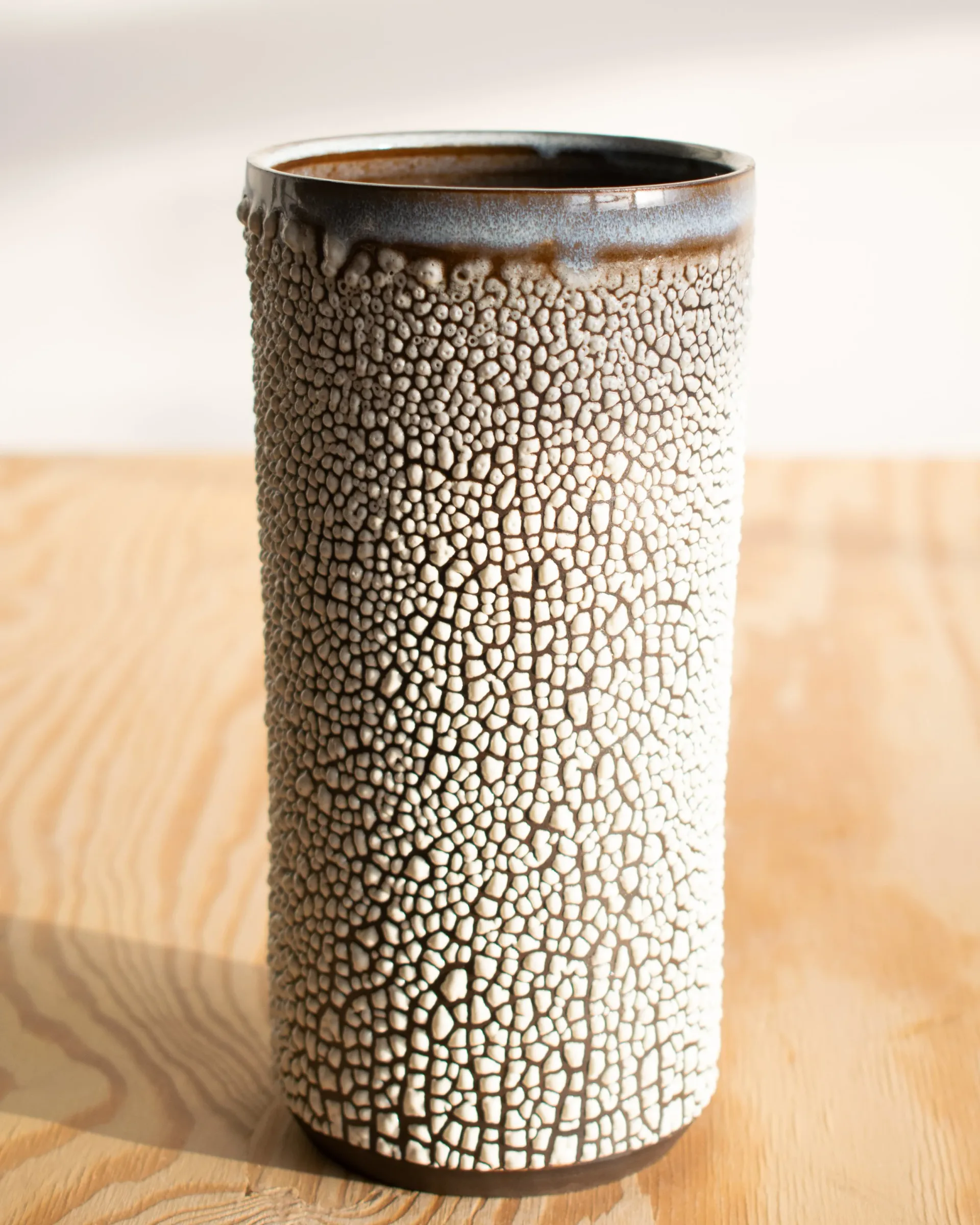 Cylindrical ceramic vessel with textured white crackle surface and dark glazed rim, displayed on natural wood by Estelle's Atelier.