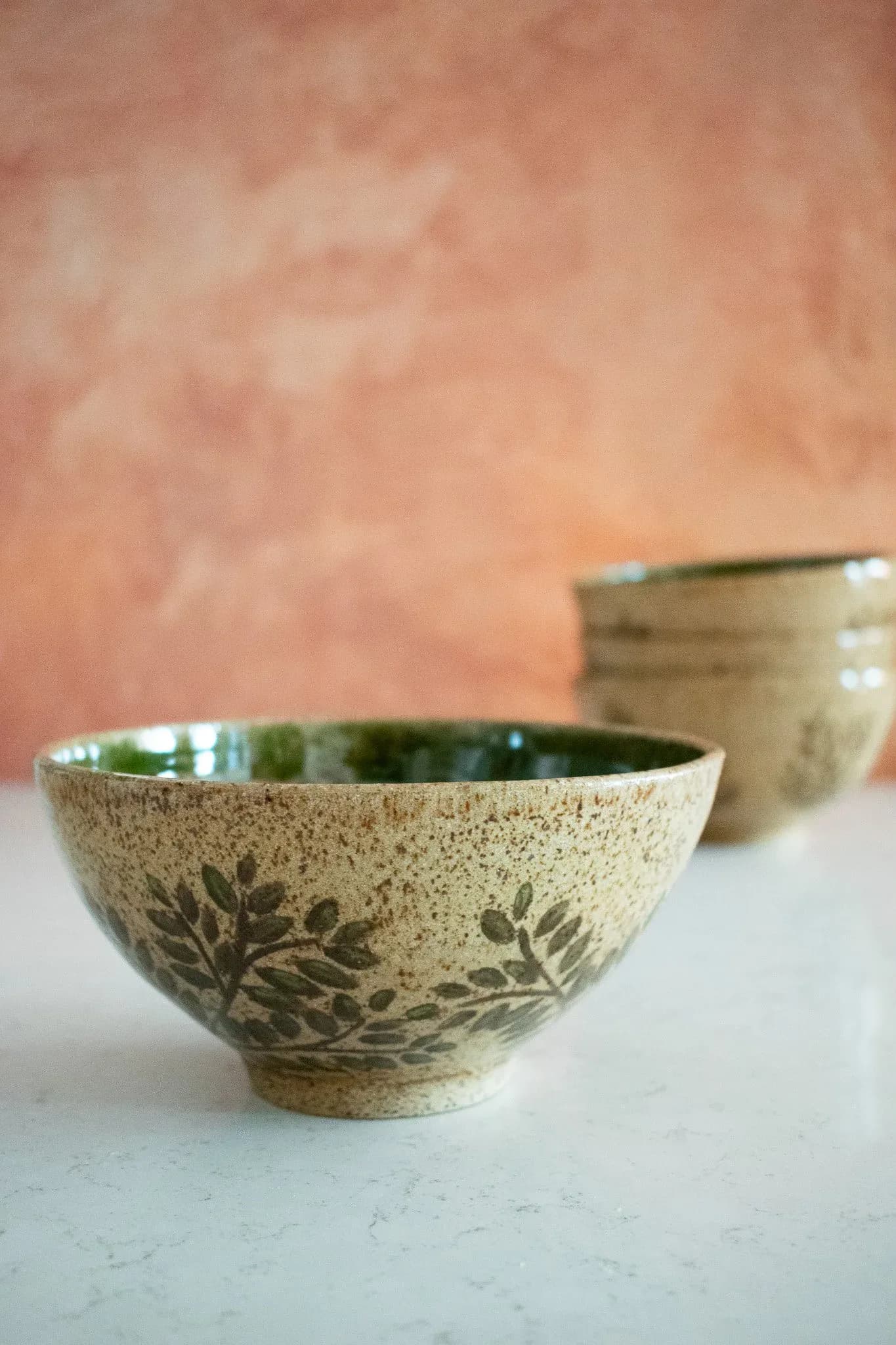 Two handcrafted ceramic bowls with textured sand-colored glaze and leaf impressions, featuring glossy green interiors, displayed against a soft pink textured wall by Estelle's Atelier.