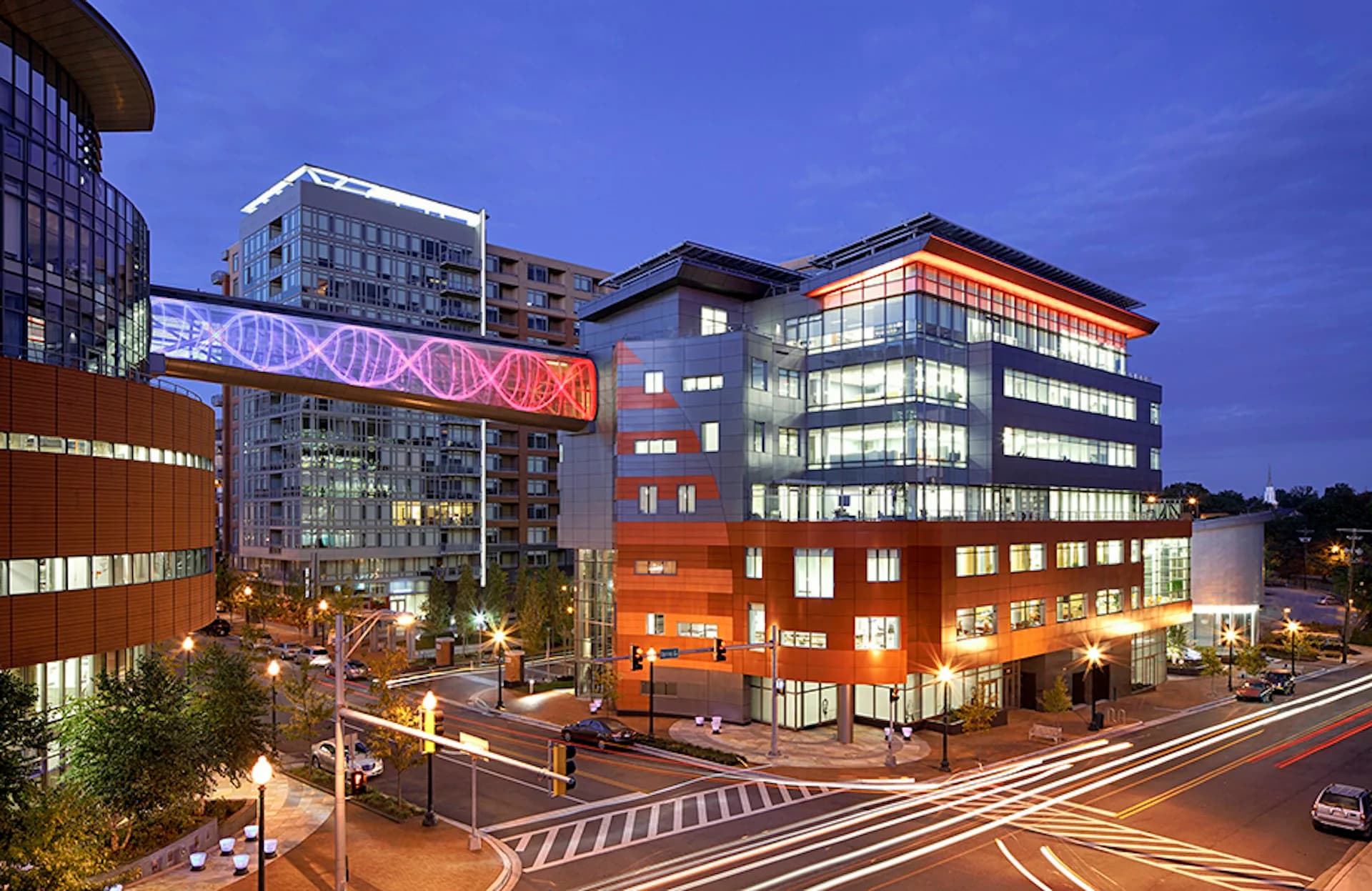 Twilight urban streetscape featuring a vibrant mixed-use development with illuminated orange and blue buildings, modern glass facades, and pedestrian plaza by EwingCole.