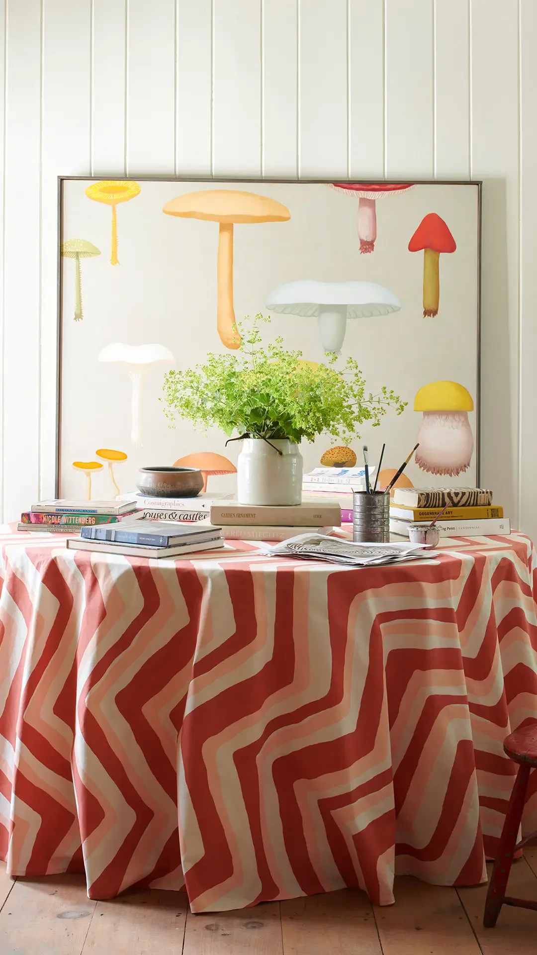 Whimsical styled tabletop display featuring striped red and cream tablecloth, potted green plant, stacked books, art supplies, and illustrated mushroom backdrop by F. Schumacher & Co.