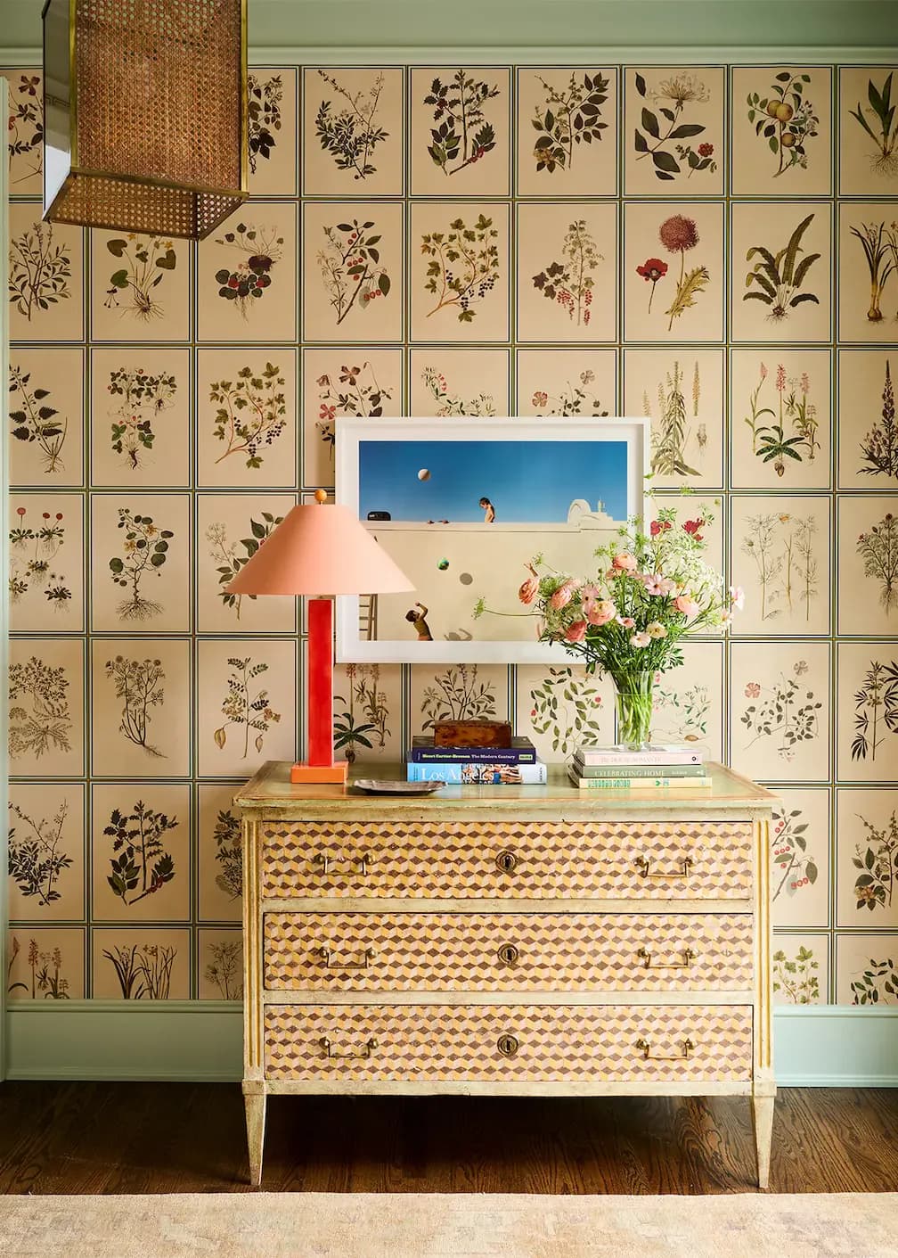 Sage green interior featuring a botanical wallpaper wall with botanical prints, topped with a patterned dresser, coral lamp, and floral arrangement by F. Schumacher & Co.