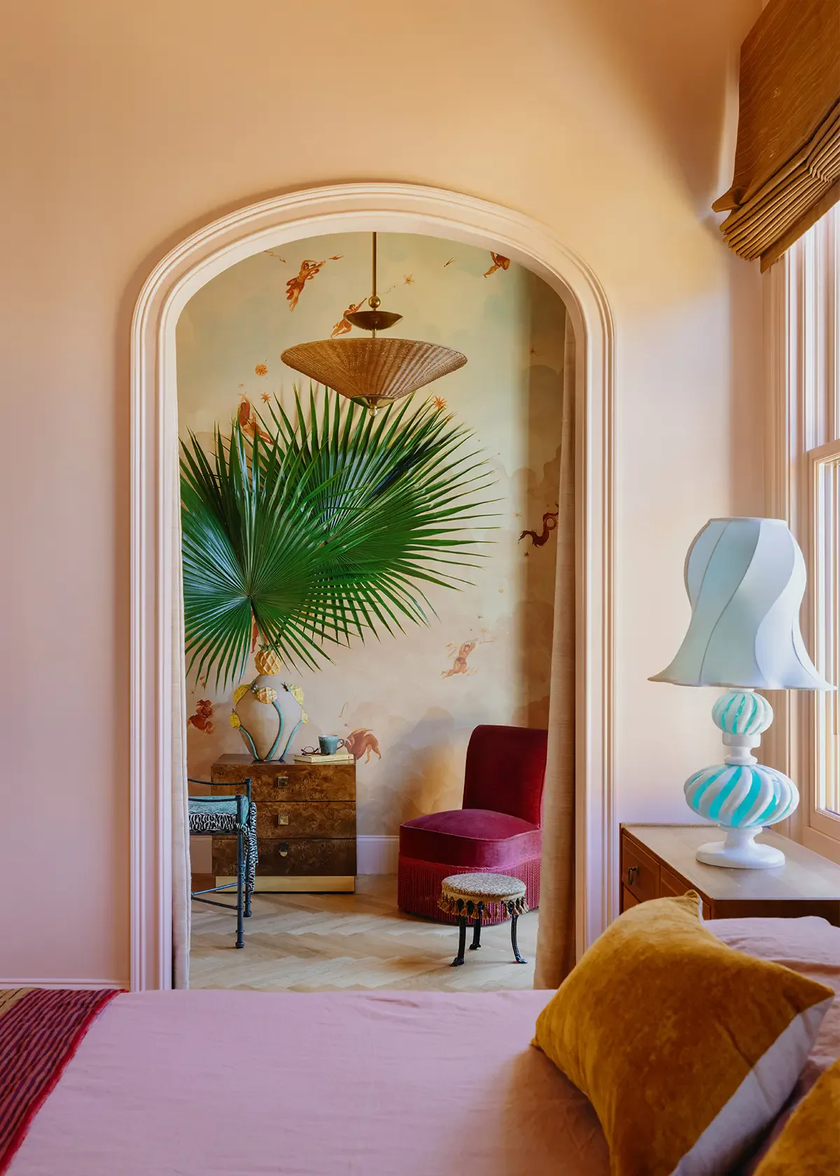 Arched doorway opening to a sunlit room with palm plants, burgundy velvet chair, wooden furniture, and decorative brass pendant, showcasing warm eclectic interiors by F. Schumacher & Co.