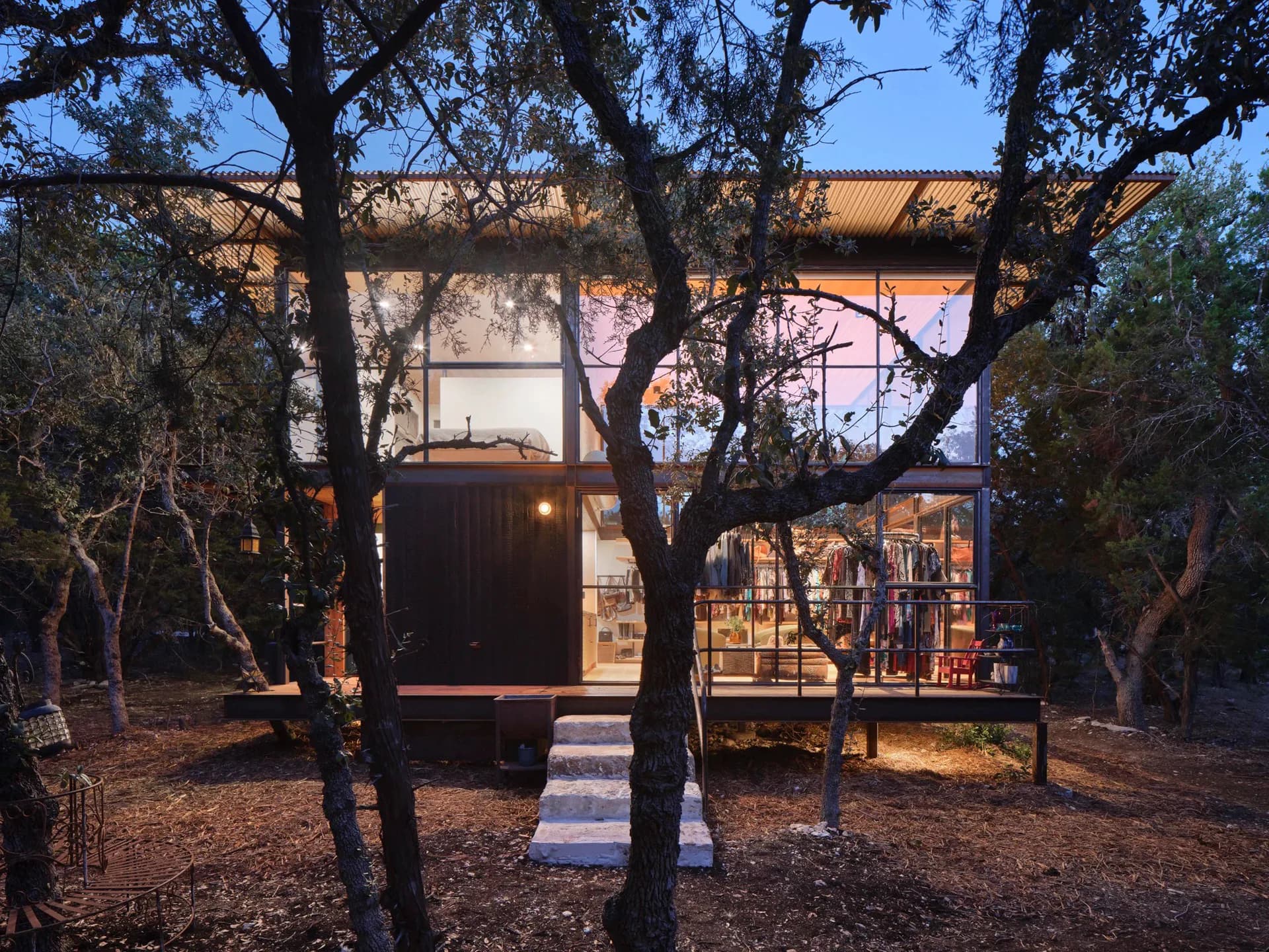 Evening view of a modern pavilion with warm interior lighting framed by gnarled olive trees and natural landscape, designed by FAR Architecture.