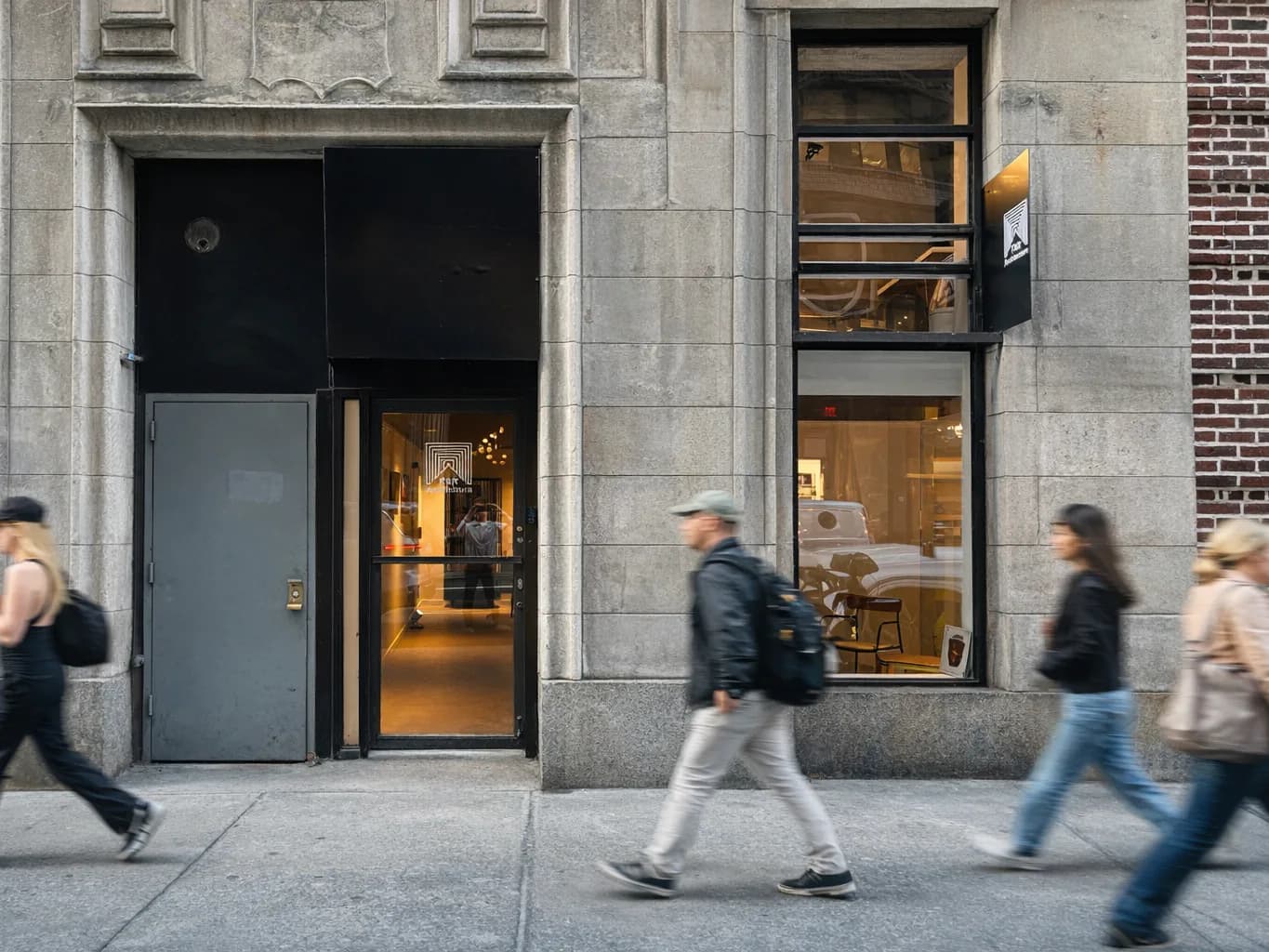 Street-level storefront with gray stone facade, glazed entry door and windows, warm interior lighting, pedestrians passing by, designed by FAR Architecture.