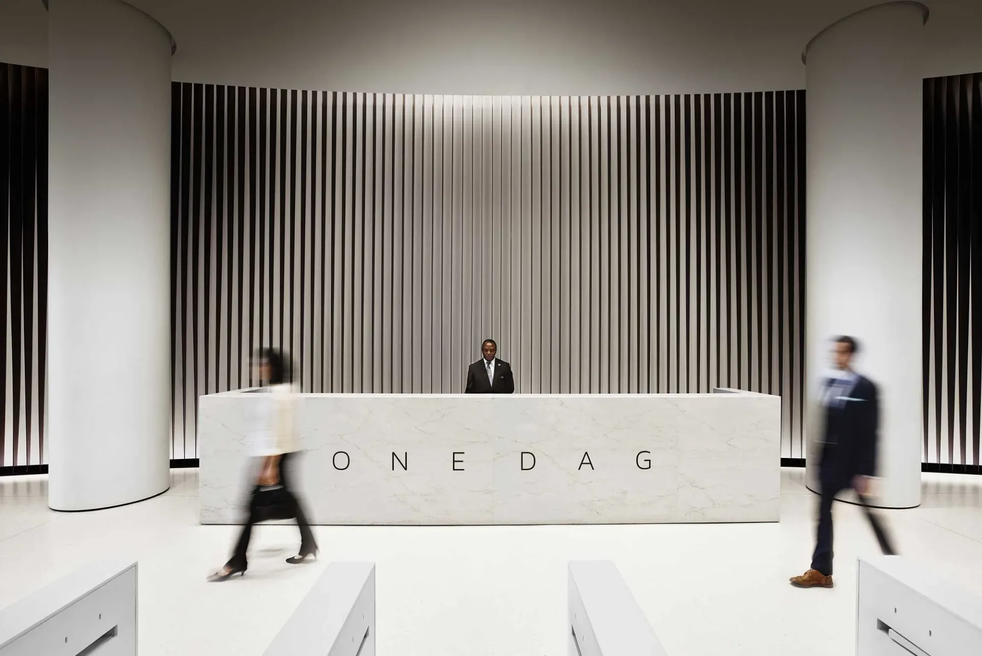 Minimalist reception desk with vertical slat wall backdrop and 'ONEDAG' lettering, featuring clean lines and monochromatic palette by Ferra Designs.