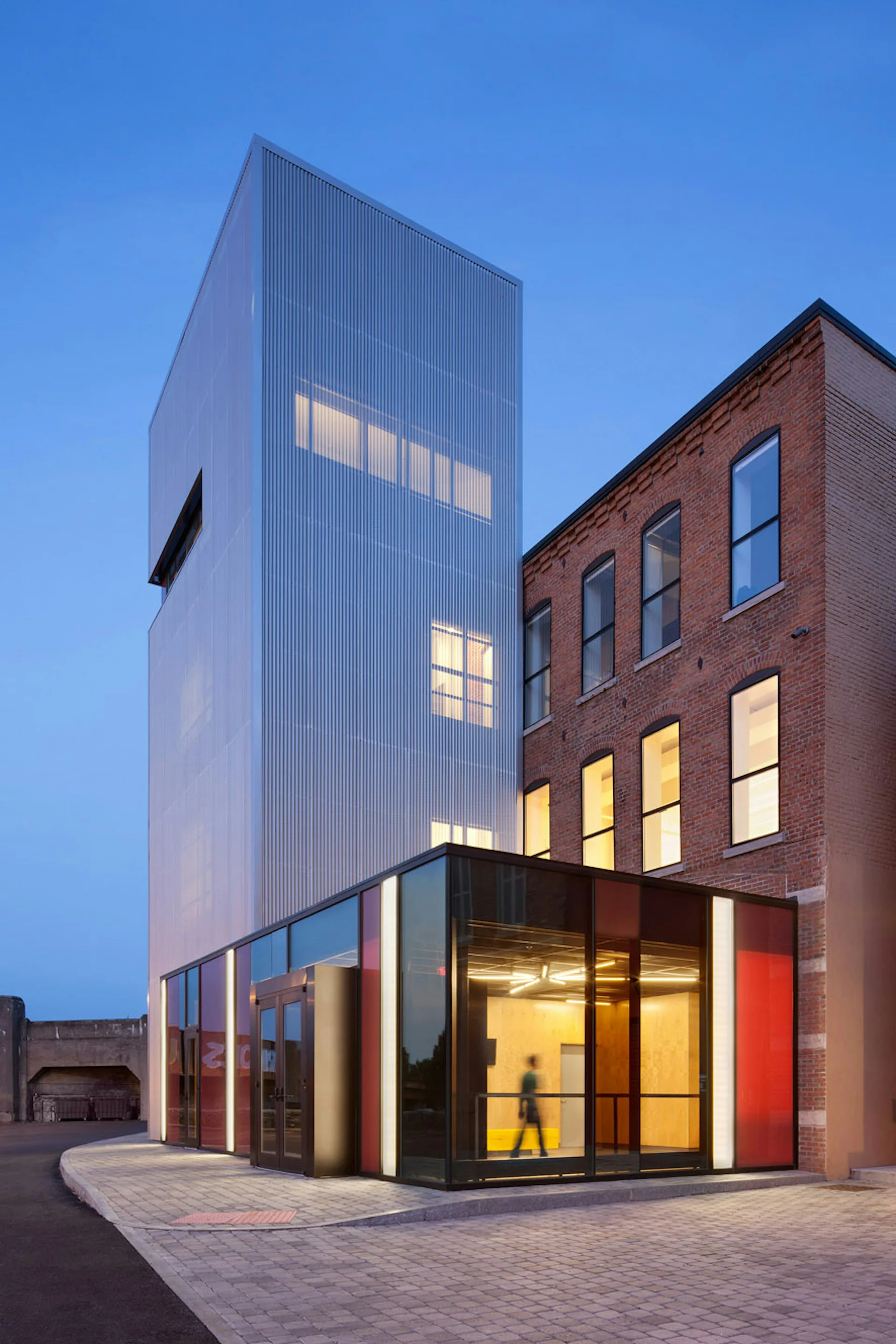 Modern mixed-use building exterior at dusk featuring a tall corrugated metal tower with illuminated windows beside a restored brick facade, designed by Fiedler Marciano Architecture.