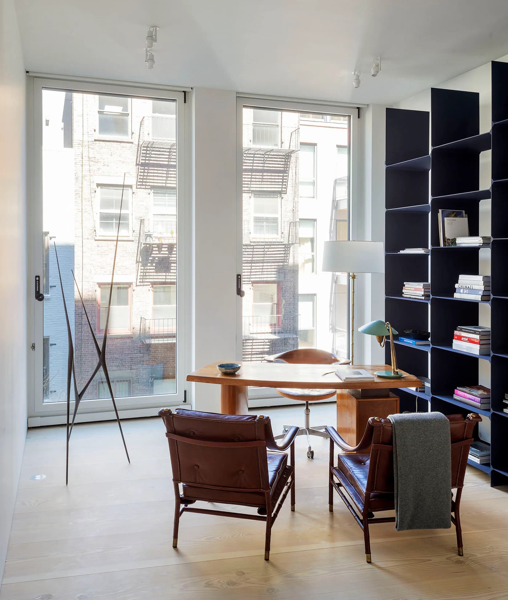 Modern office space with floor-to-ceiling windows, mid-century wooden desk, brown leather chairs, and dark blue shelving by Fiedler Marciano Architecture.
