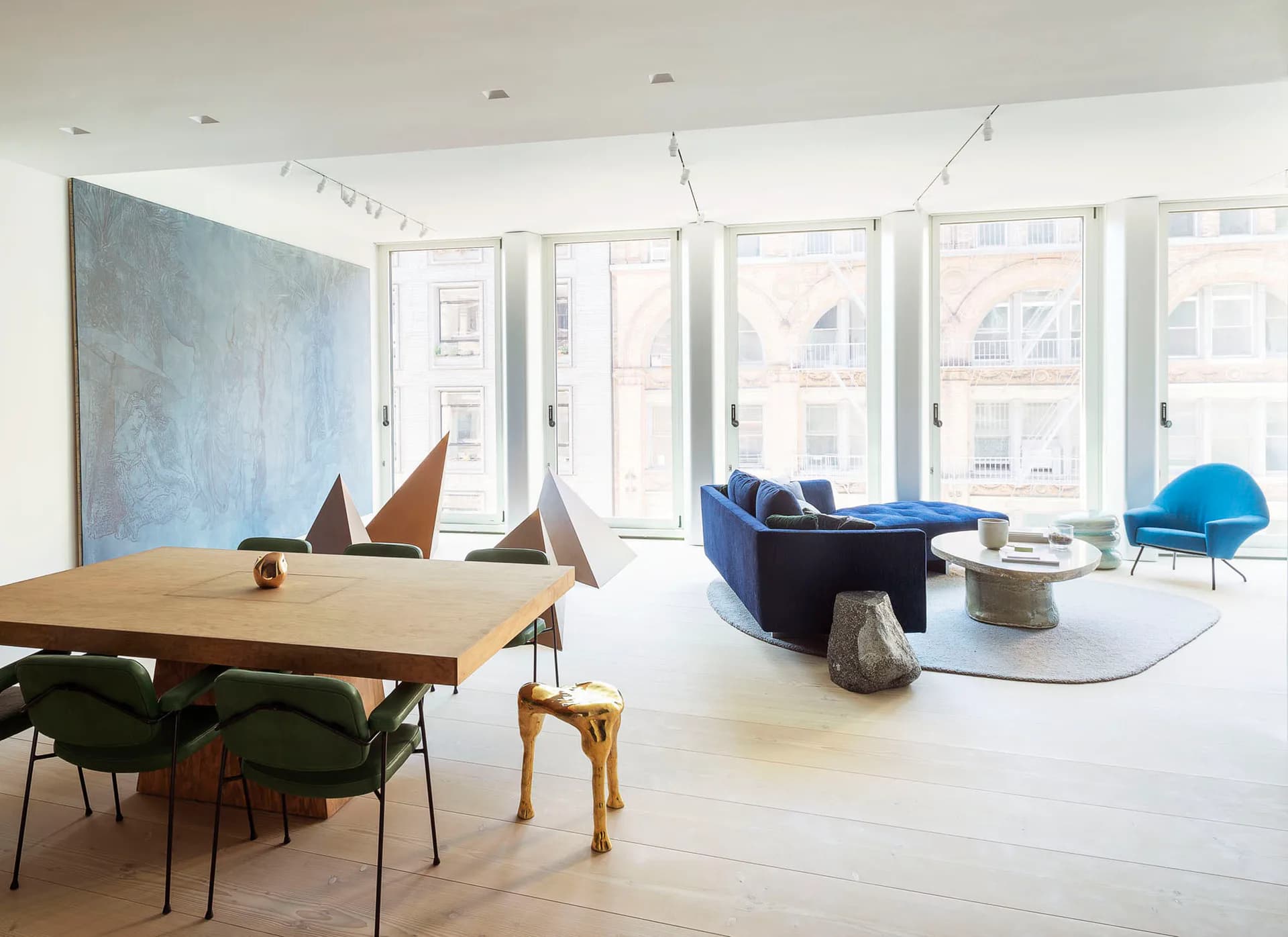 Bright modern loft with wooden dining table, blue seating, sculptural elements, and expansive windows overlooking city streets by Fiedler Marciano Architecture.
