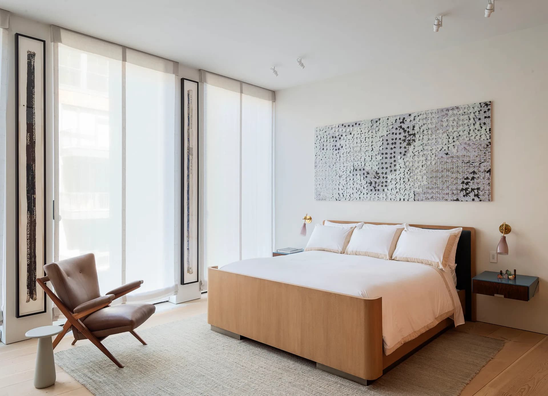 Minimalist bedroom with warm wood bed frame, floor-to-ceiling windows, leather lounge chair, and textured wall art by Fiedler Marciano Architecture.