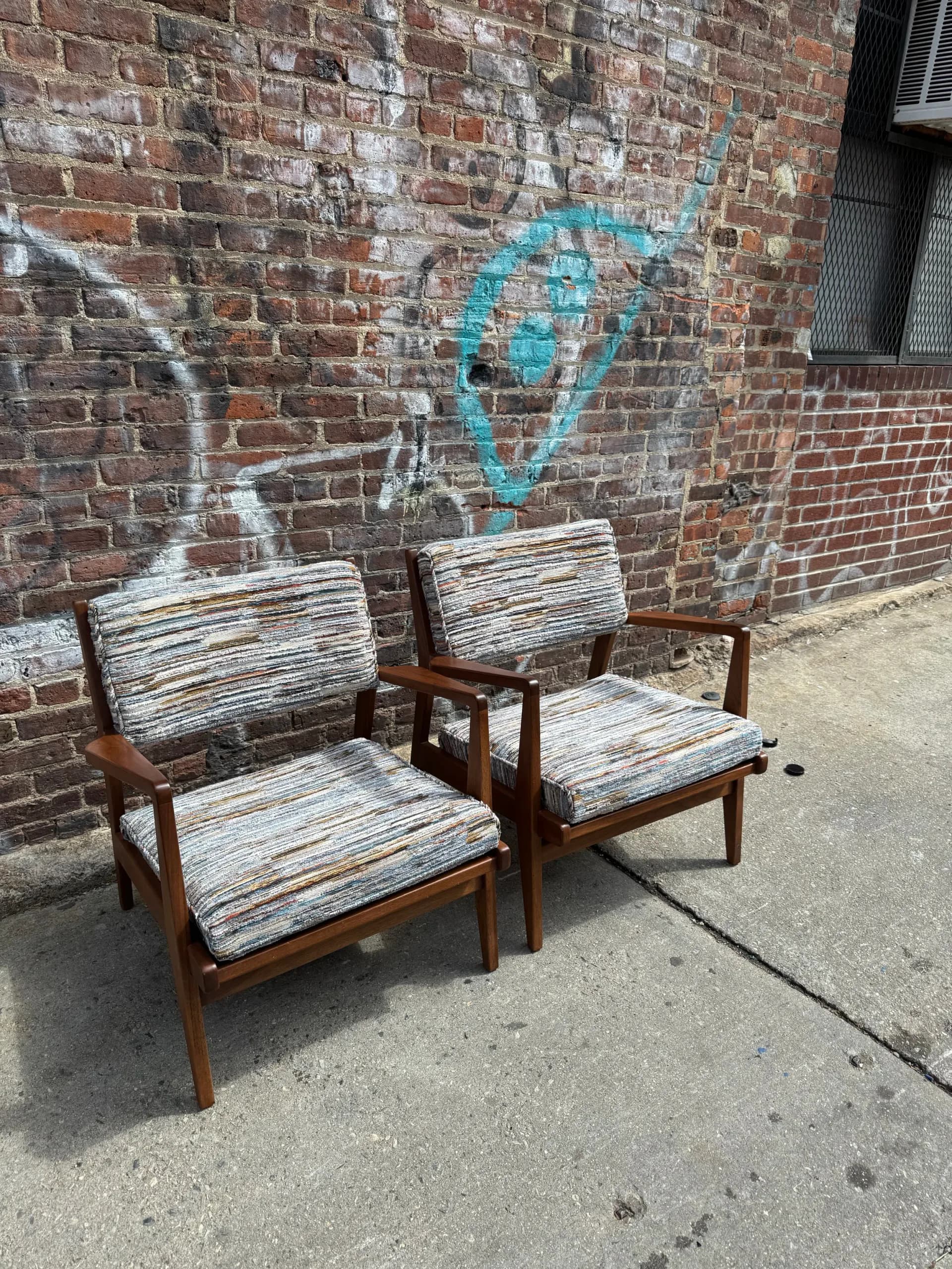 Two mid-century modern wooden chairs with striped upholstery positioned against a brick wall with blue graffiti art by Find the Thread Upholstery.