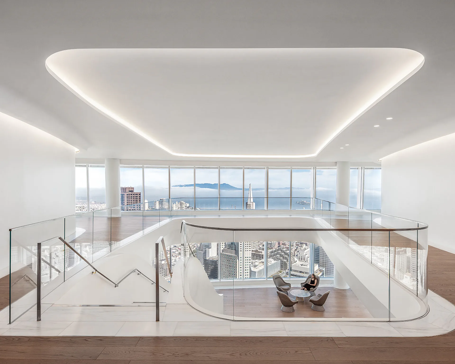 Modern high-rise interior with curved white ceiling, glass railings, and floor-to-ceiling windows overlooking a city skyline, featuring minimalist furniture and synthetic lighting by Fisher Marantz Stone.