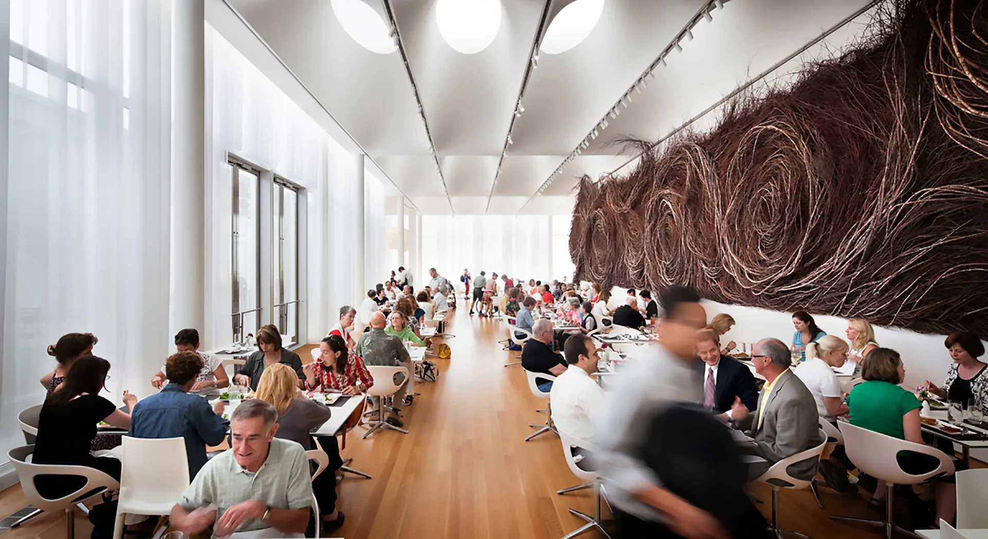 Modern cafeteria with wooden flooring, white columns, large abstract brown artwork, and numerous seated diners throughout the bright, open space by Fisher Marantz Stone.