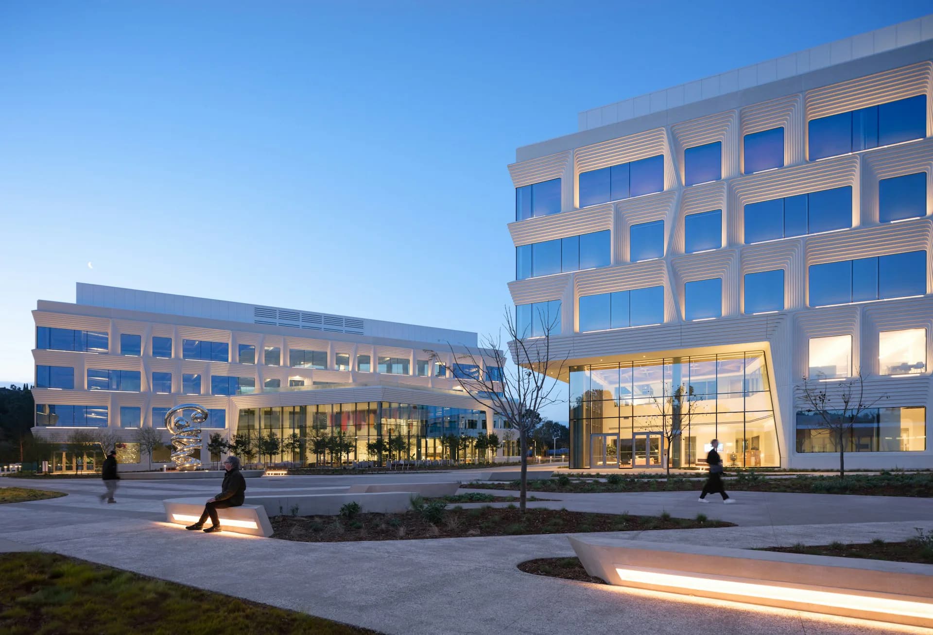 Modern institutional complex with illuminated glass-fronted lobbies, clean geometric facades, and plaza with integrated landscape lighting by Flad Architects.