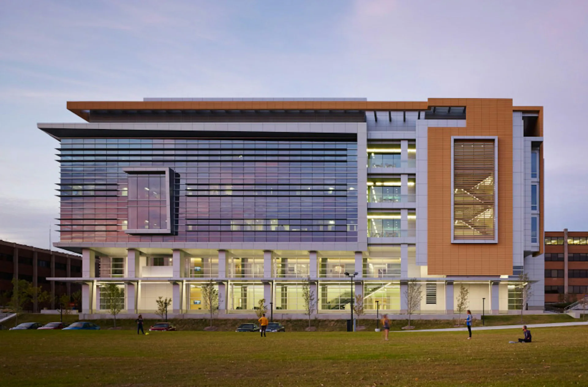 Modern institutional building with glass and terracotta facades, linear stacked windows, and illuminated ground-floor gallery spaces overlooking lawn by Flad Architects.
