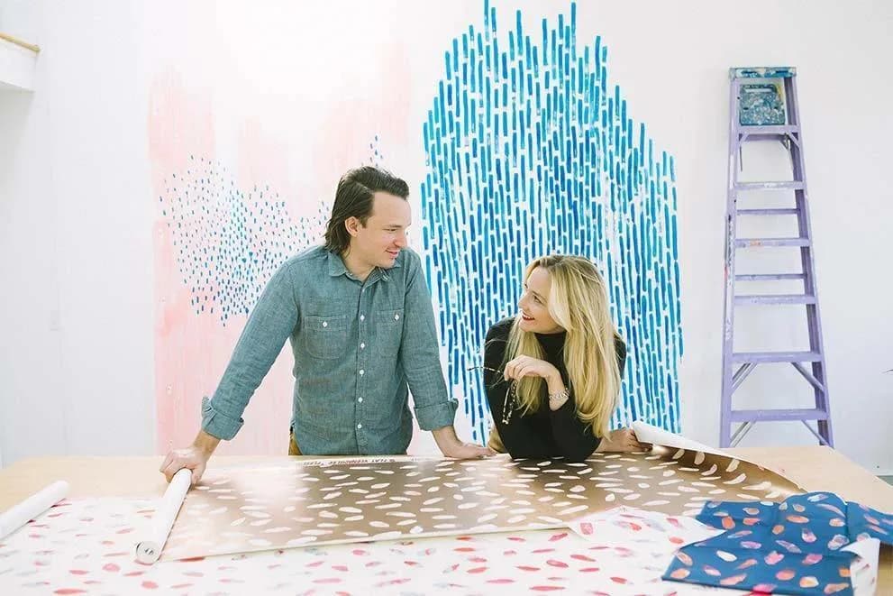 Two people collaborate on a wall installation featuring geometric patterns in blue, pink, and neutral tones with a ladder visible, by Flat Vernacular.