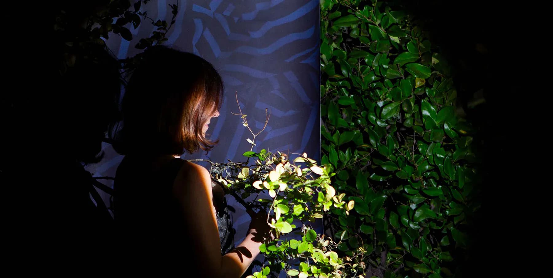 Woman interacting with illuminated green living wall installation against dark backdrop with blue patterned element by Flat Vernacular.