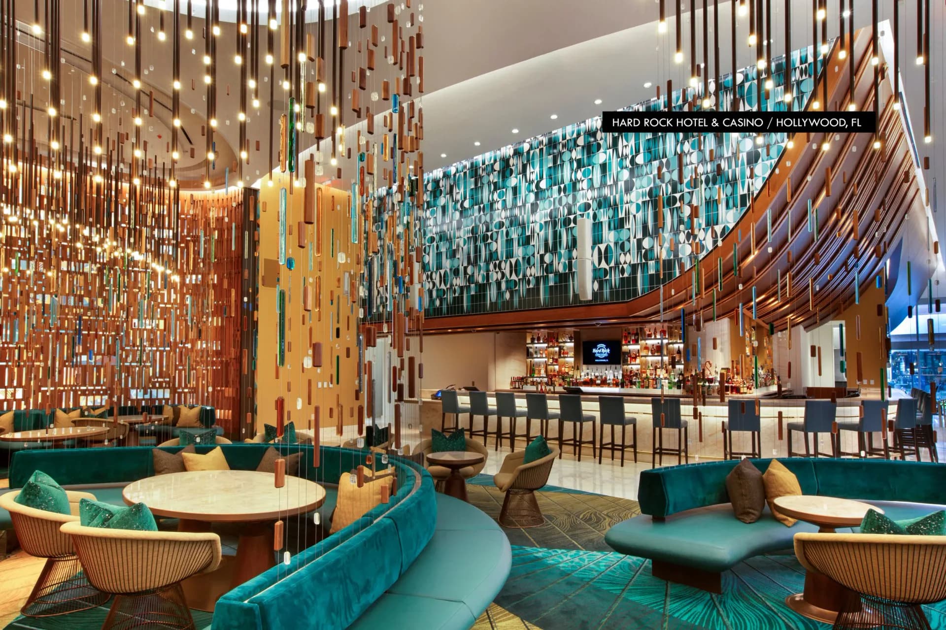 Hotel lobby with turquoise and gold color scheme, featuring hanging glass pendant installations, curved furniture, and geometric wall patterns by Focus Lighting.