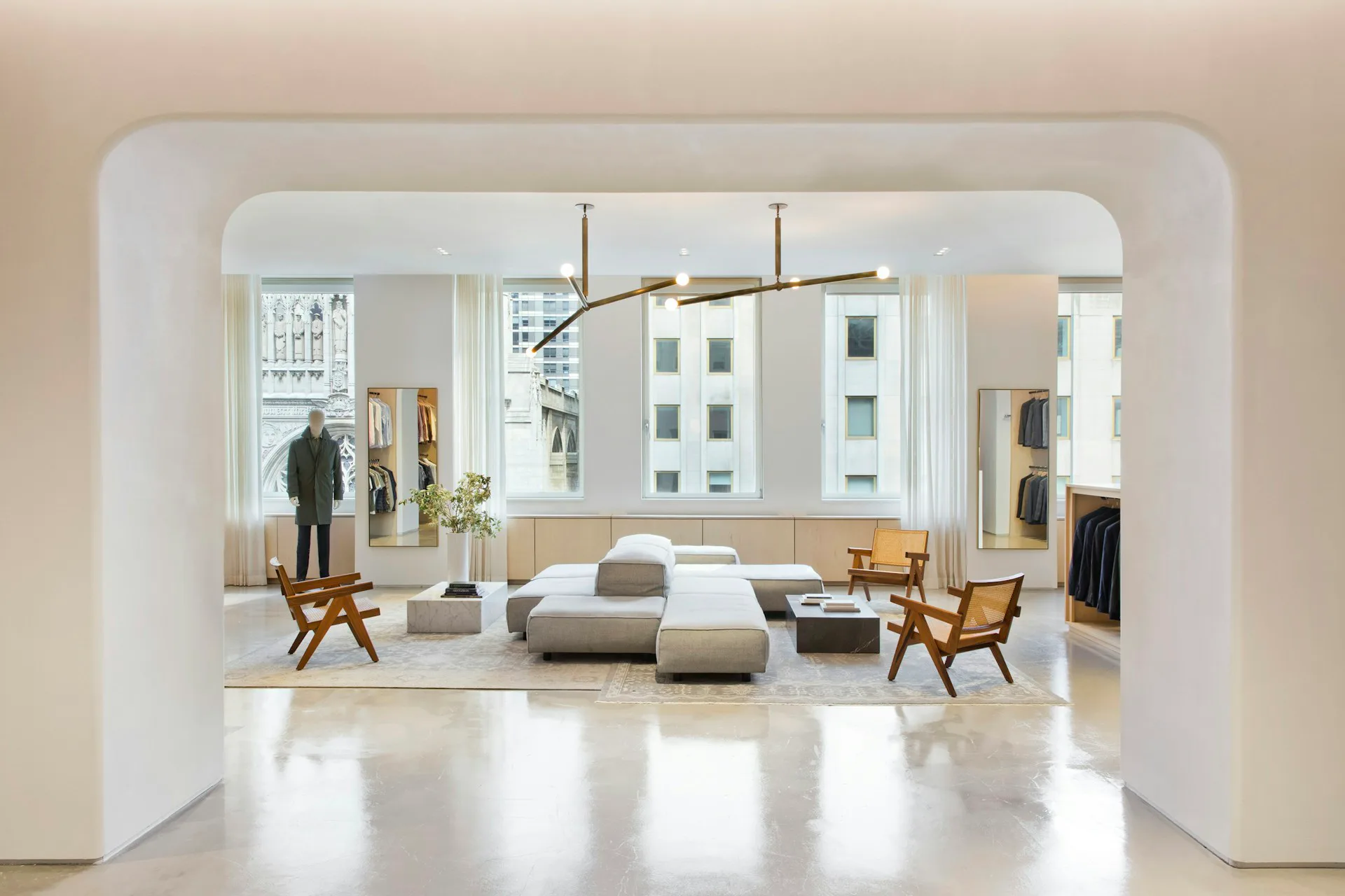 A minimalist interior showroom with cream walls, polished concrete floors, and symmetrically arranged furniture including tan upholstered seating, wooden chairs, and clothing displays, designed by Fogarty Finger.