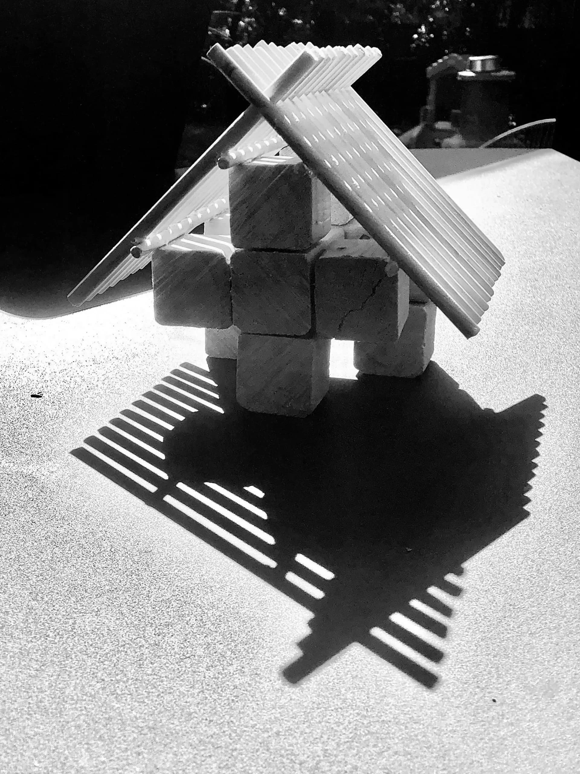Black and white photograph of a sculptural wooden installation with stacked cubic forms and crossed beam elements, casting geometric shadows on a light surface, by FORRM.