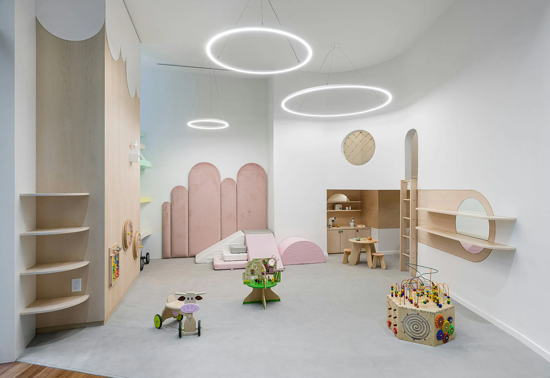 Pastel children's playroom with pink curved furniture, circular pendant lights, wooden shelving, and toy displays by FORRM.