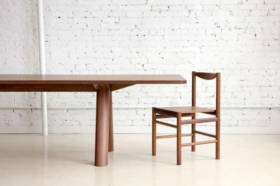Minimalist wooden dining table with tapered legs and matching chair featuring woven seat, set against white brick wall by Fort Standard.