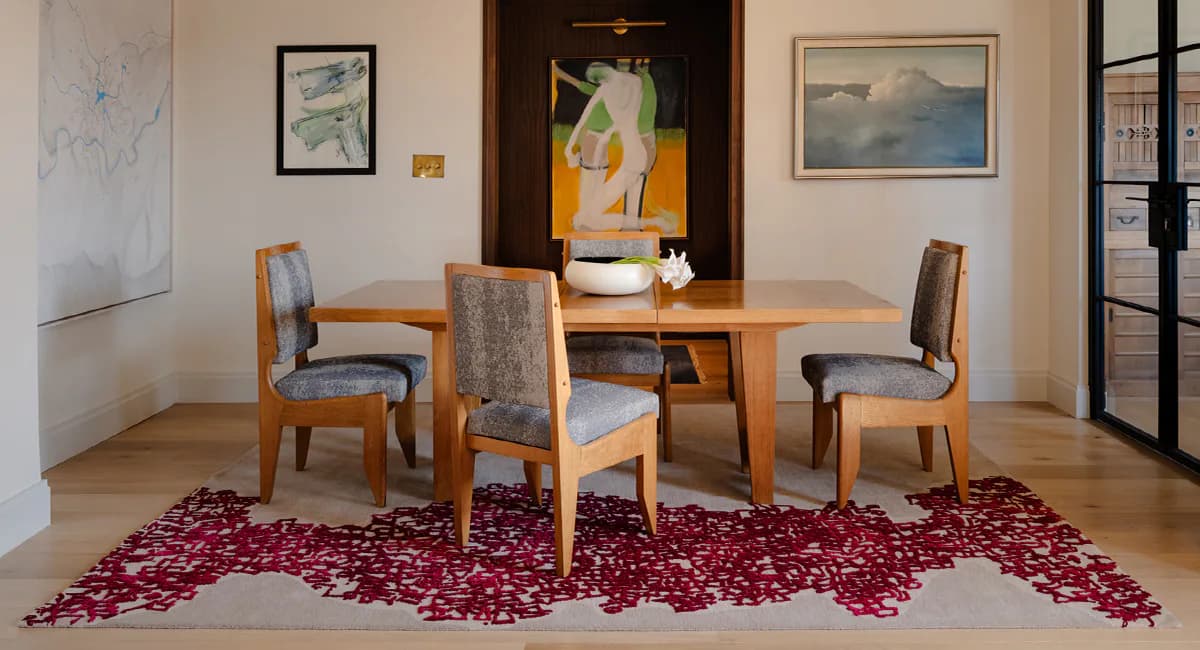 A dining room interior features a wooden table with upholstered chairs set on a burgundy patterned rug, framed by gallery walls and modern fixtures by Fort Street Studio.
