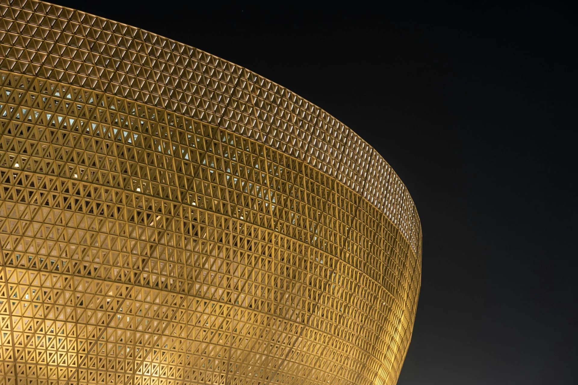 Curved golden mesh facade with geometric pattern illuminated against dark night sky, architectural detail by Foster + Partners.