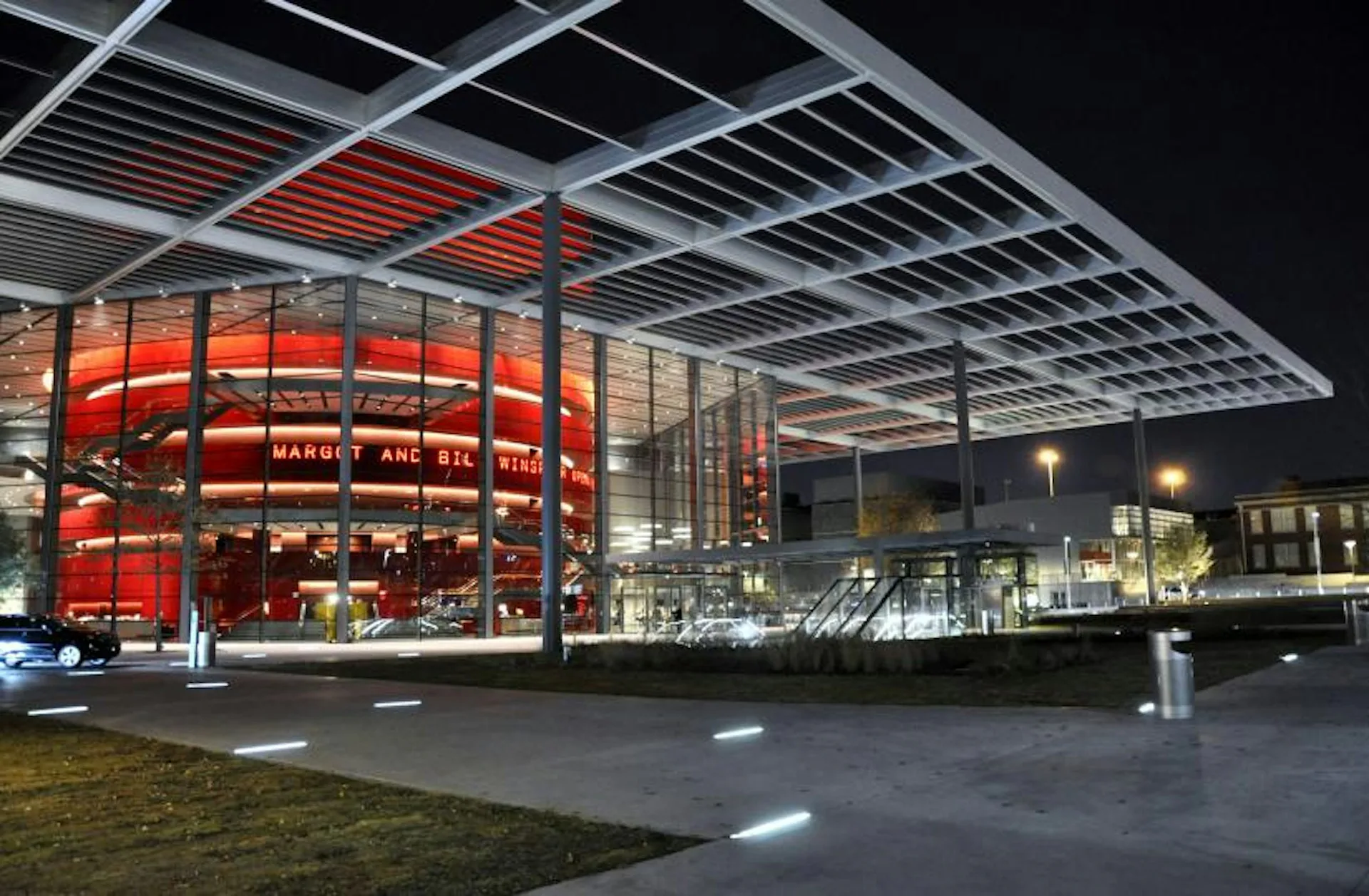 Night view of a modern building with a distinctive red-illuminated circular glass façade and angular steel canopy overhead, designed by Foster + Partners.