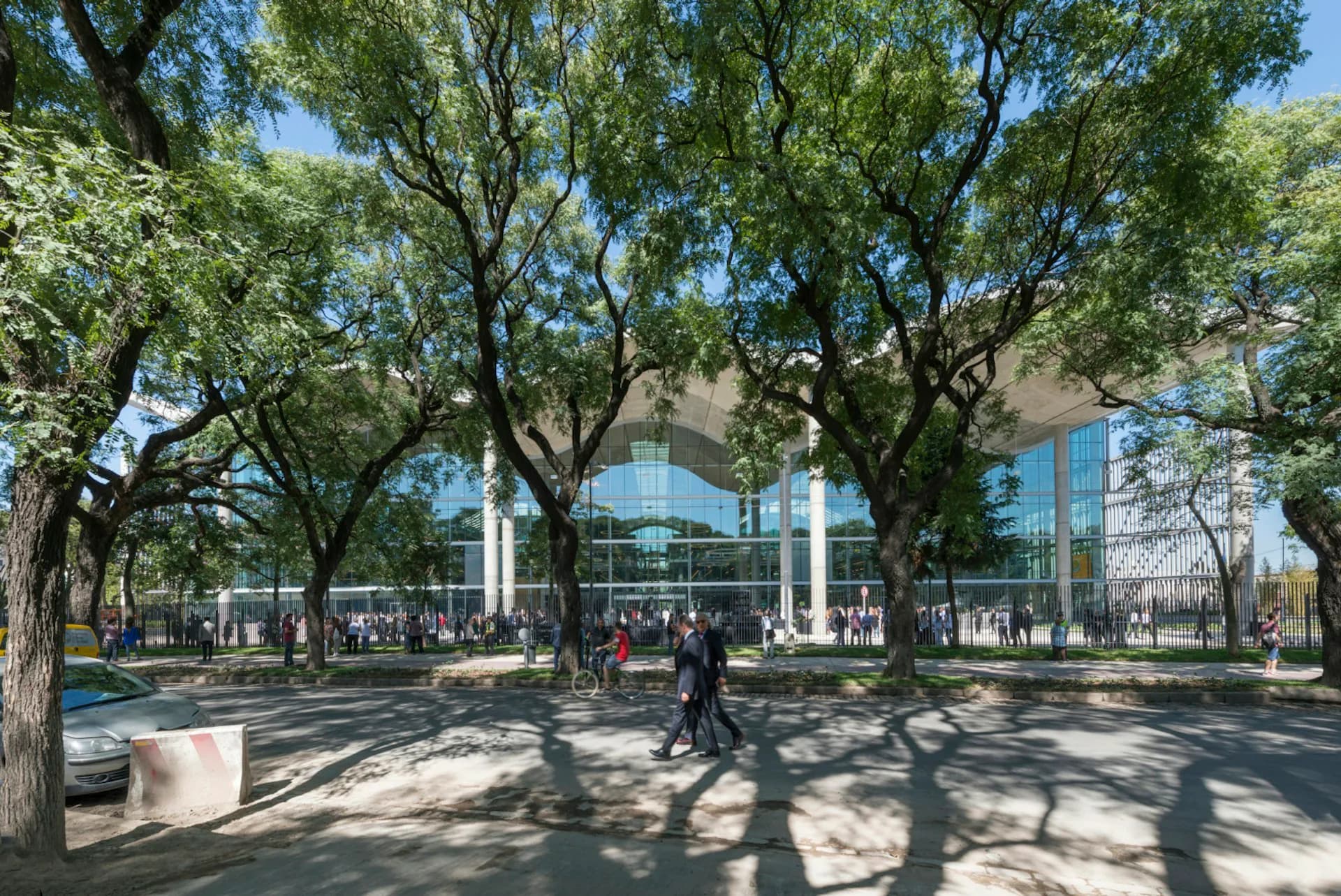 Public plaza with mature trees, modern glass building facade, and pedestrians enjoying a sunlit courtyard space designed by Foster + Partners.