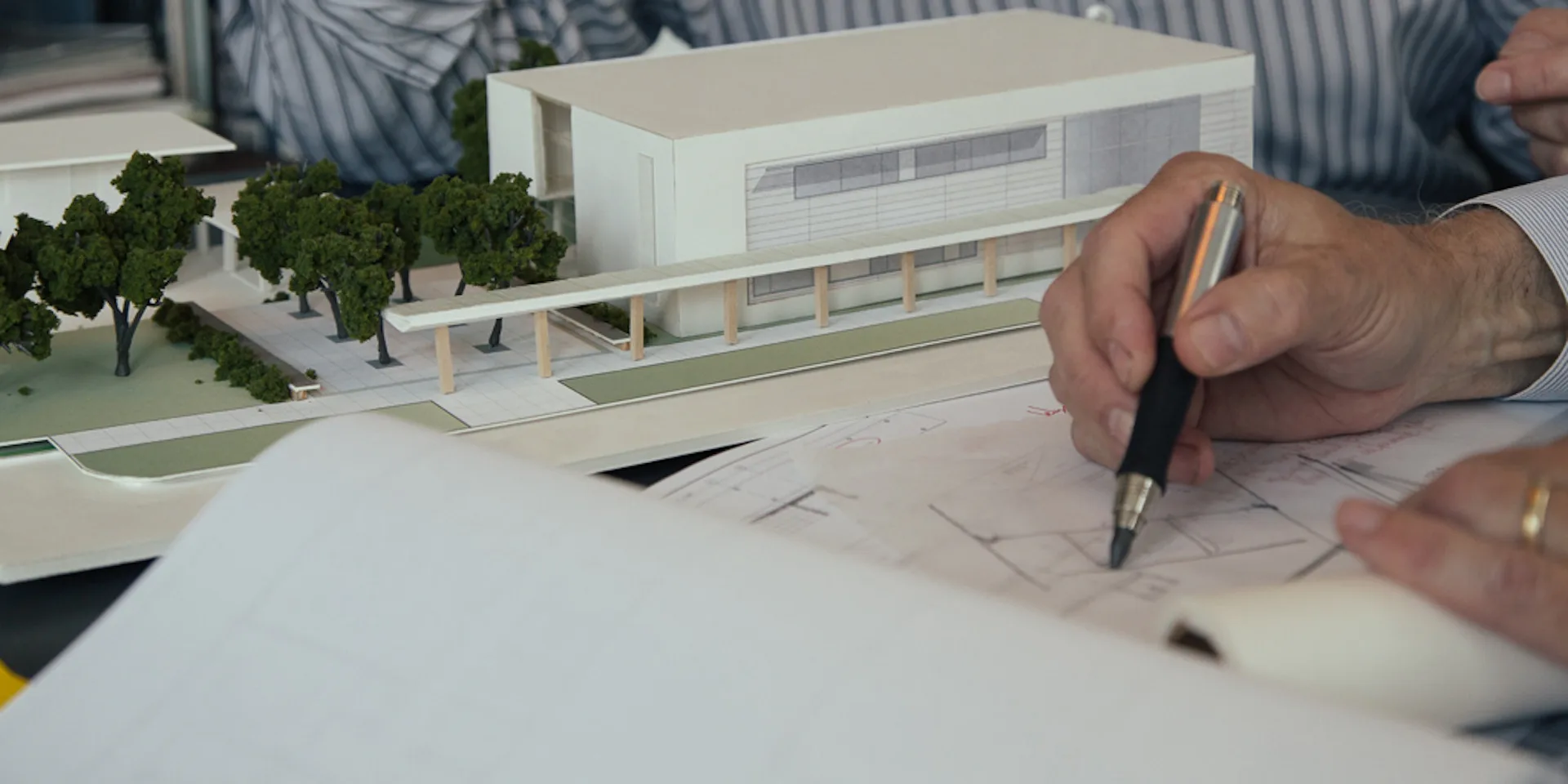 Architect's hand sketching design details on paper with an architectural model and landscape elements visible on the workspace by Fradkin Cooper.