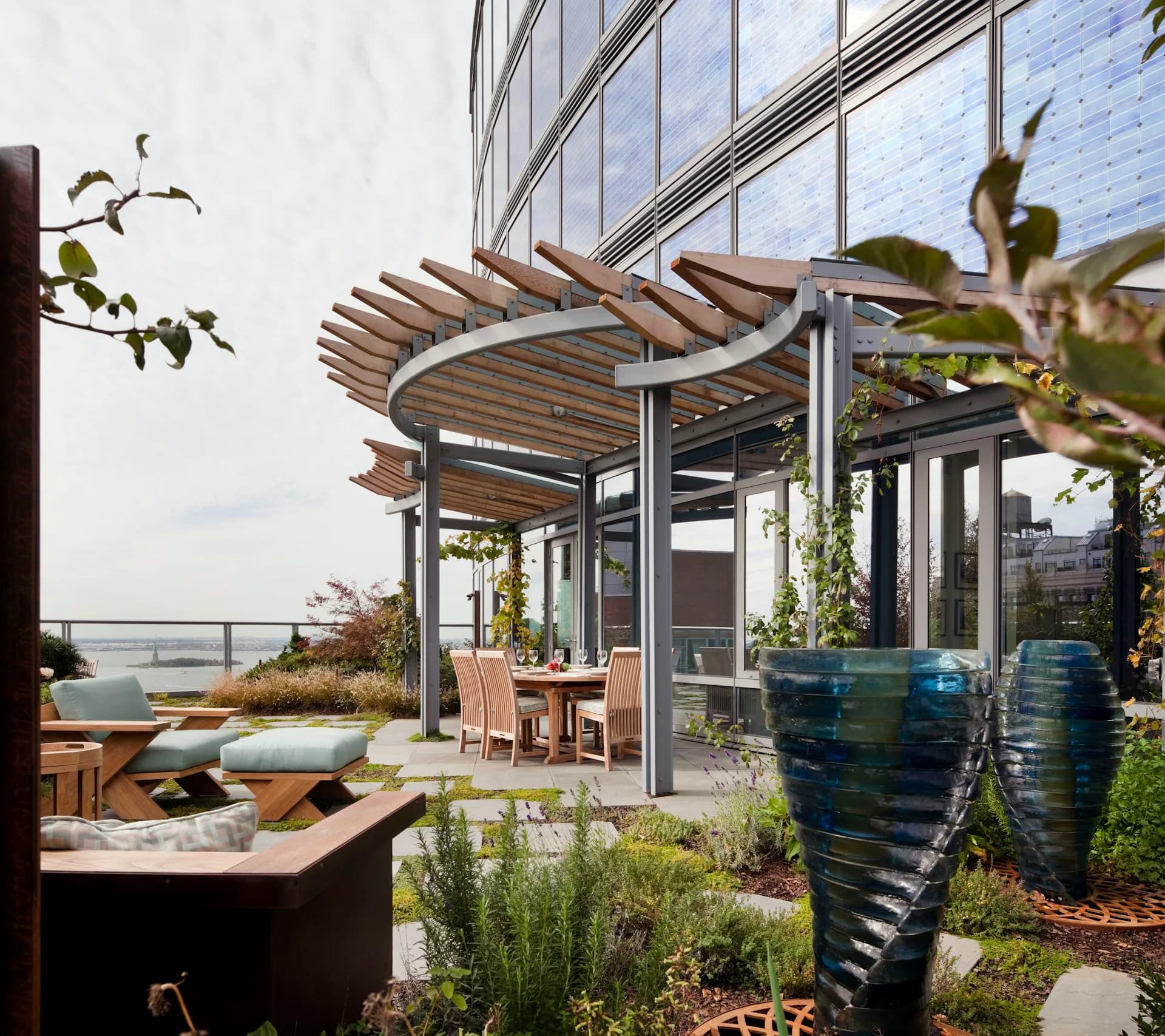 Modern rooftop terrace with curved wooden pergola, glass sliding doors, planted urns, and water views by Fradkin Cooper.