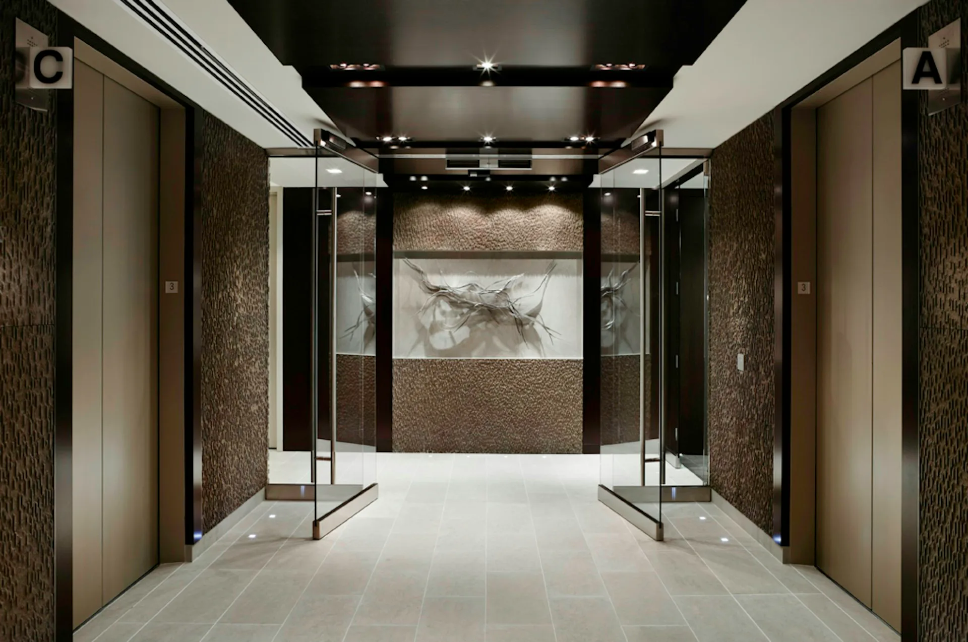 Modern luxury bathroom corridor with dual walk-in showers, marble flooring, warm wood paneling, and recessed ceiling lights by Fradkin Cooper.