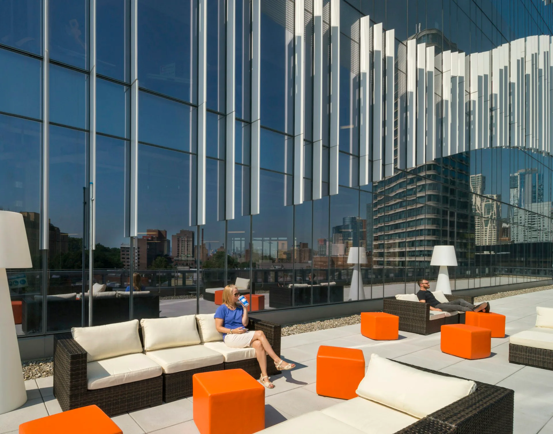 Modern rooftop terrace with orange poufs, cream sectional sofa, and panoramic city views framed by mirrored glass walls by Francis Cauffman.