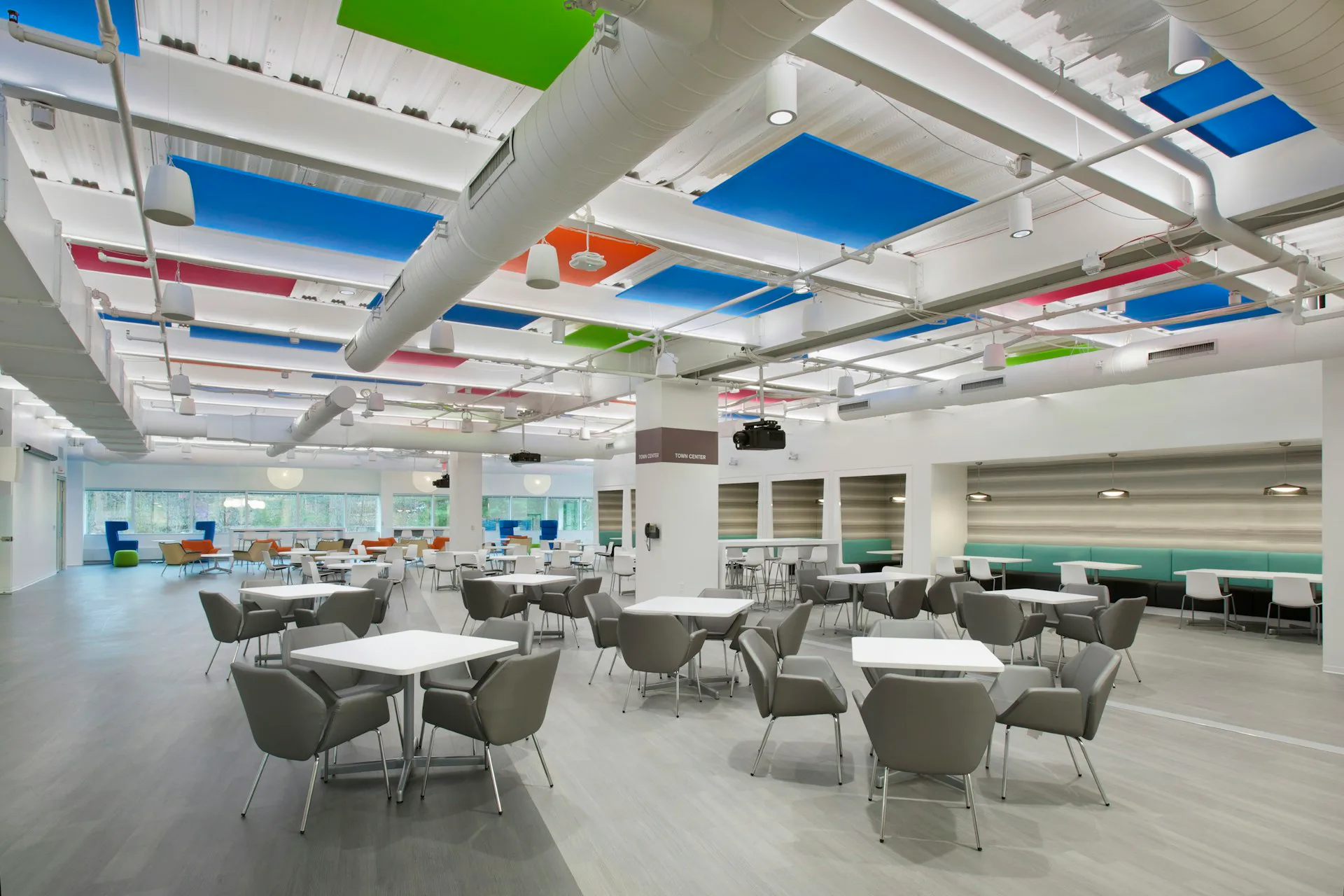Modern open office with colorful ceiling panels in blue, green, and orange, round tables with gray chairs, and collaborative workspace design by Francis Cauffman.