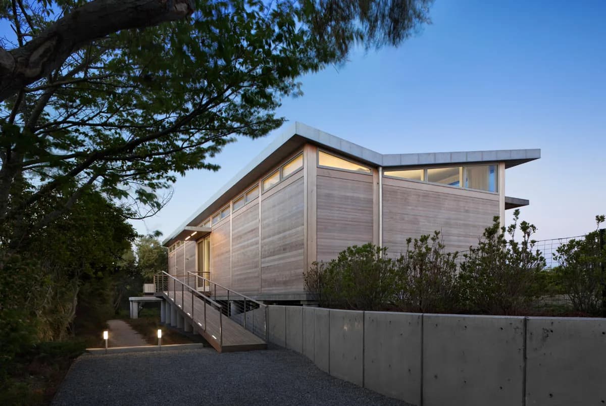 Modern elevated residential structure with horizontal wood siding and expansive glazing nestled among mature trees, featuring concrete base and metal railings by Francois de Menil Architect, PC.