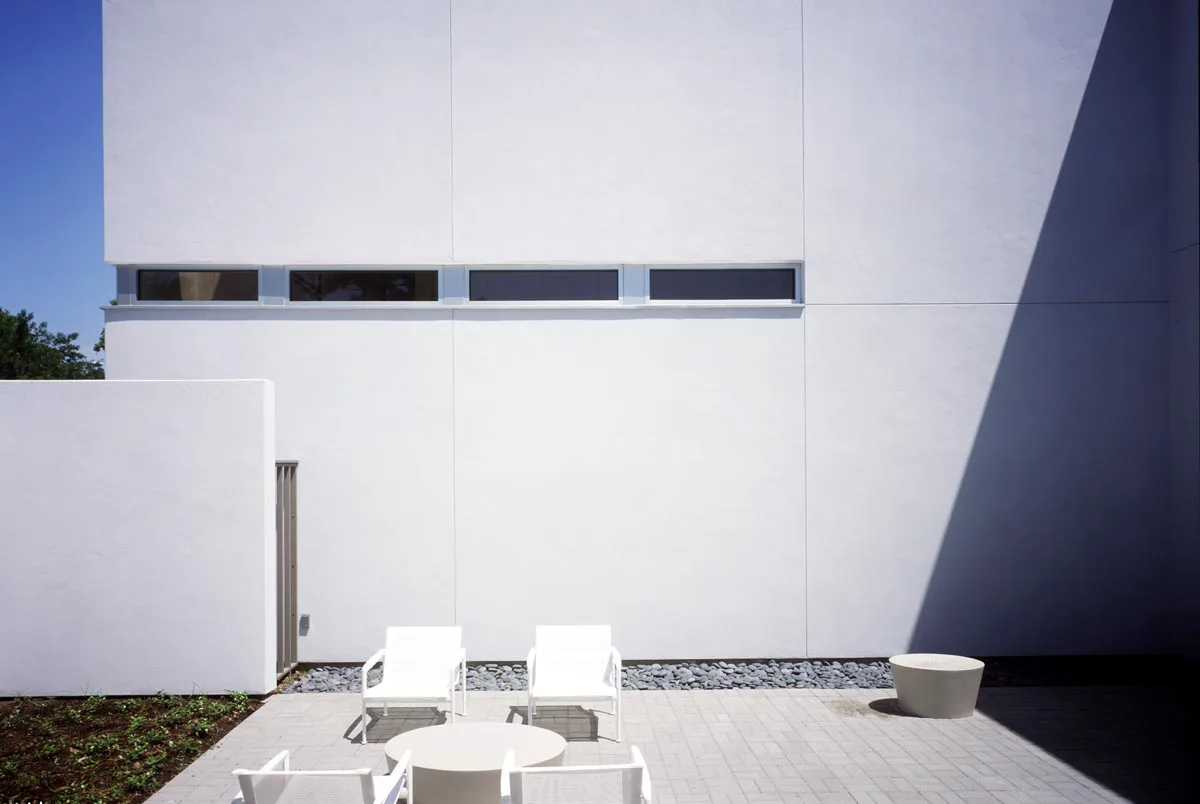 Minimalist white modernist building exterior with horizontal window strip and clean patio featuring concrete flooring and white seating by Francois de Menil Architect, PC.