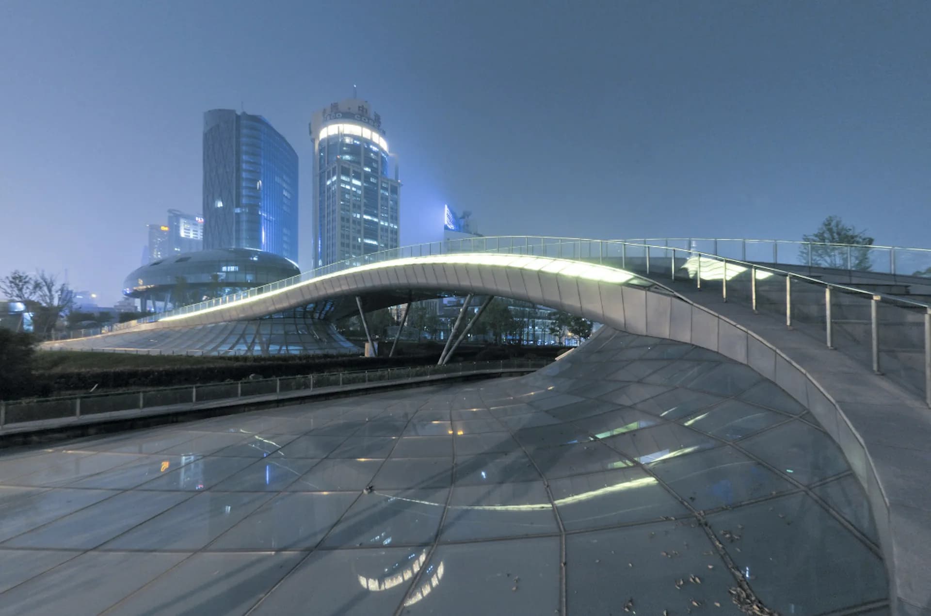 Modern curved pedestrian bridge with sleek white steel design spanning over wet pavement, flanked by contemporary glass and metal high-rise towers under clear blue sky by Frank Repas Architecture.